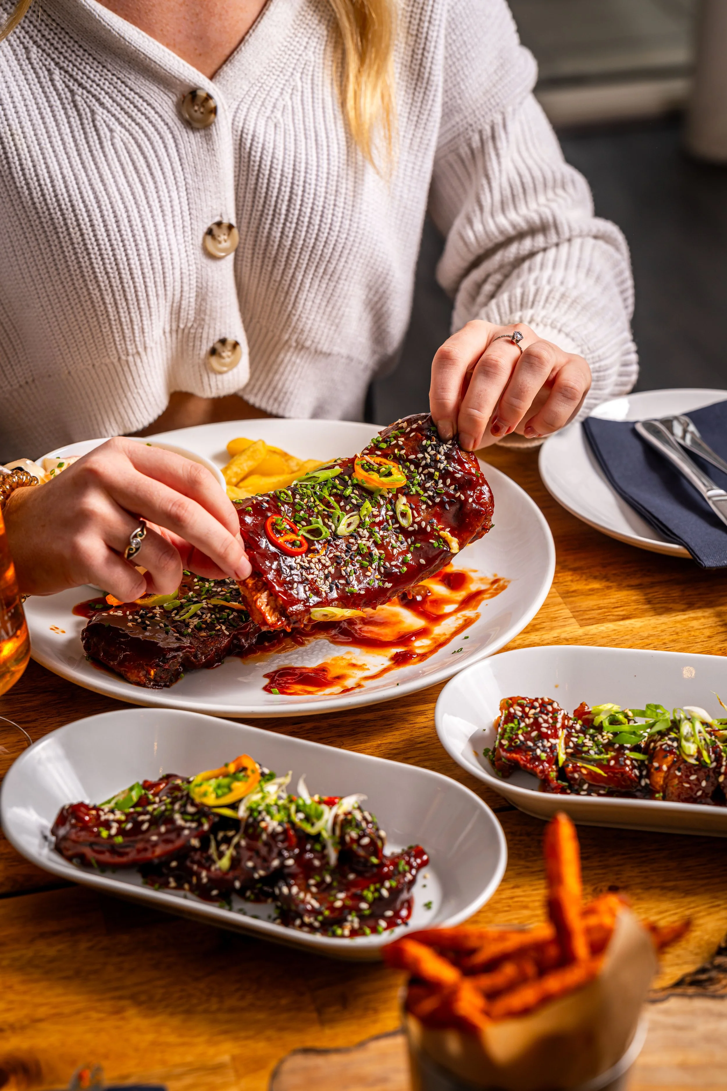 Person in a beige sweater eating grilled ribs topped with sesame seeds and sliced green onions at a wooden table with dishes of ribs and fries.