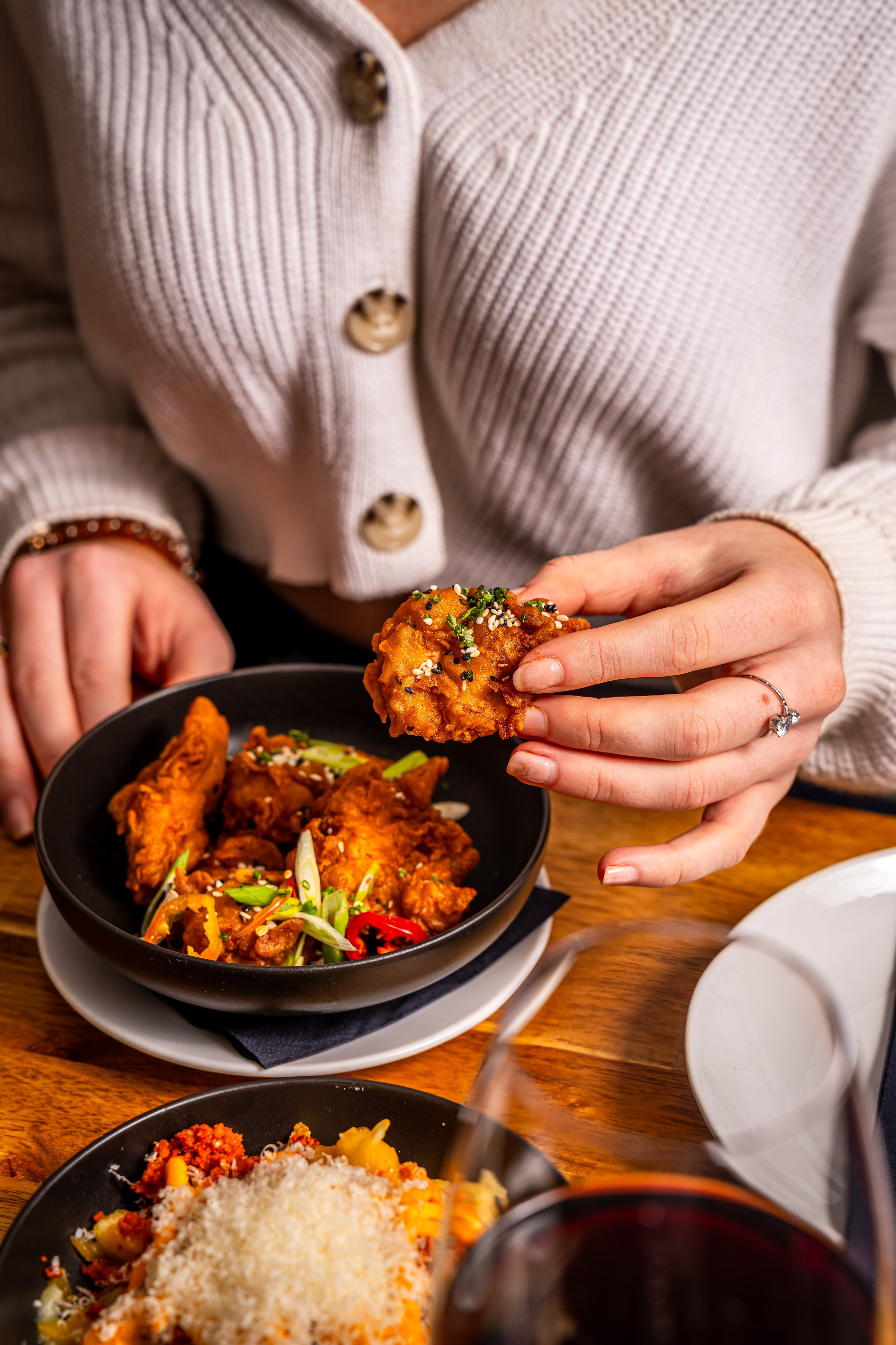 Person in a beige cardigan holding a piece of fried chicken above a black bowl of fried chicken garnished with green onions and sesame seeds, with another bowl of pasta topped with grated cheese and red sauce nearby.