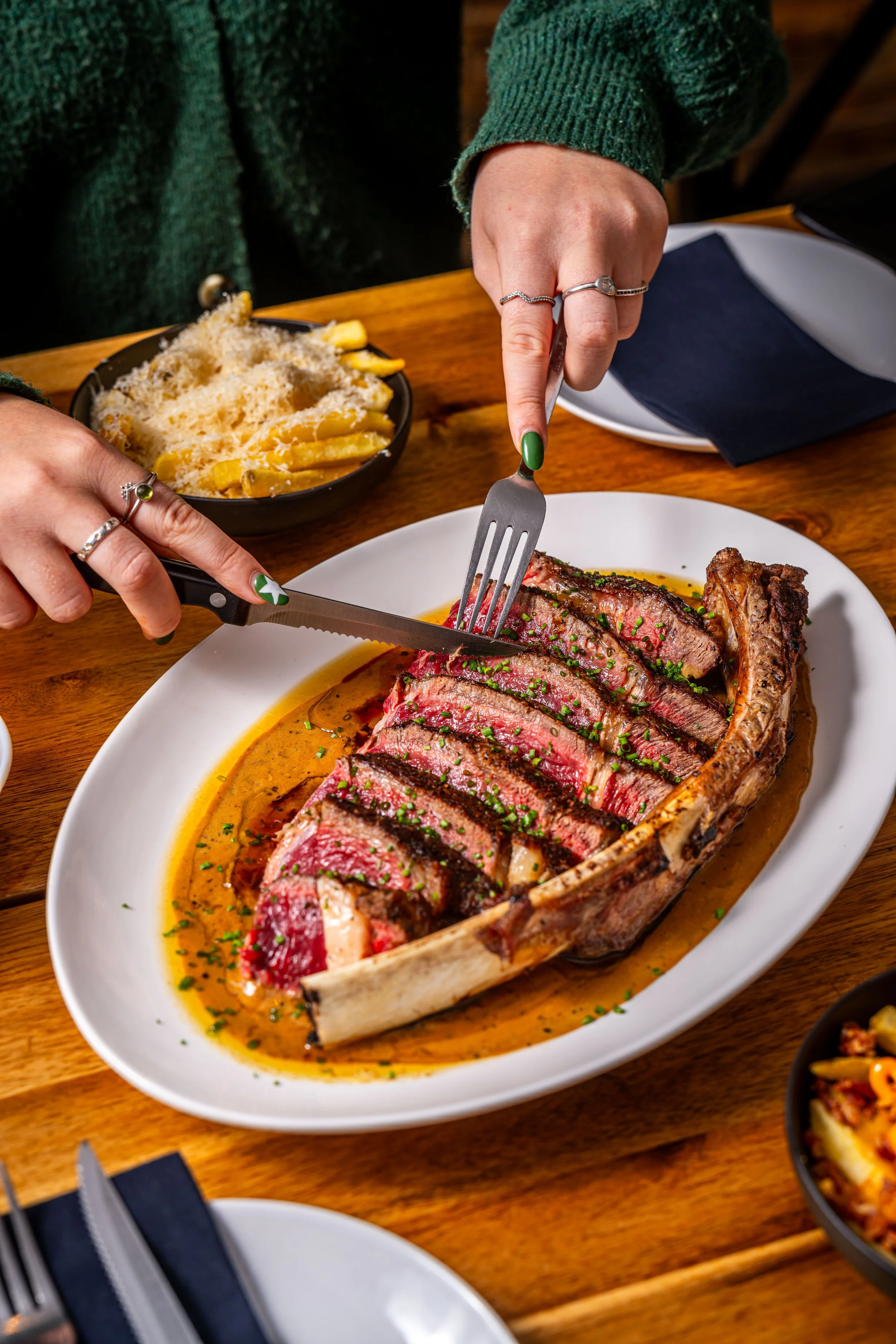 Person cutting into a cooked, rare steak on a white oval plate, with sides of pasta and salad on a wooden table.