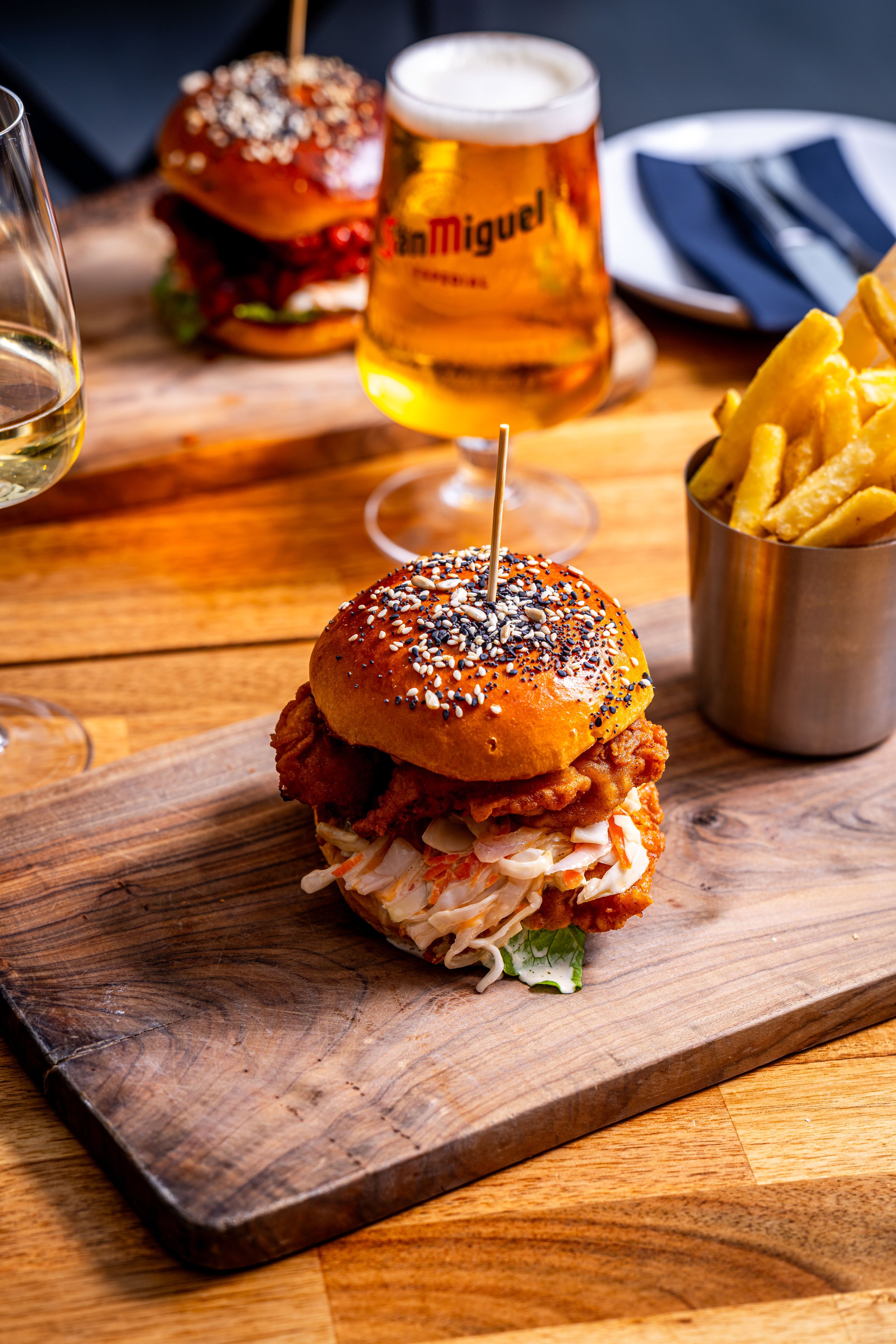 A fried chicken sandwich with coleslaw on a sesame seed bun served on a wooden cutting board, with a side of French fries in a metal cup, a glass of beer, a plate with a napkin and cutlery in the background.