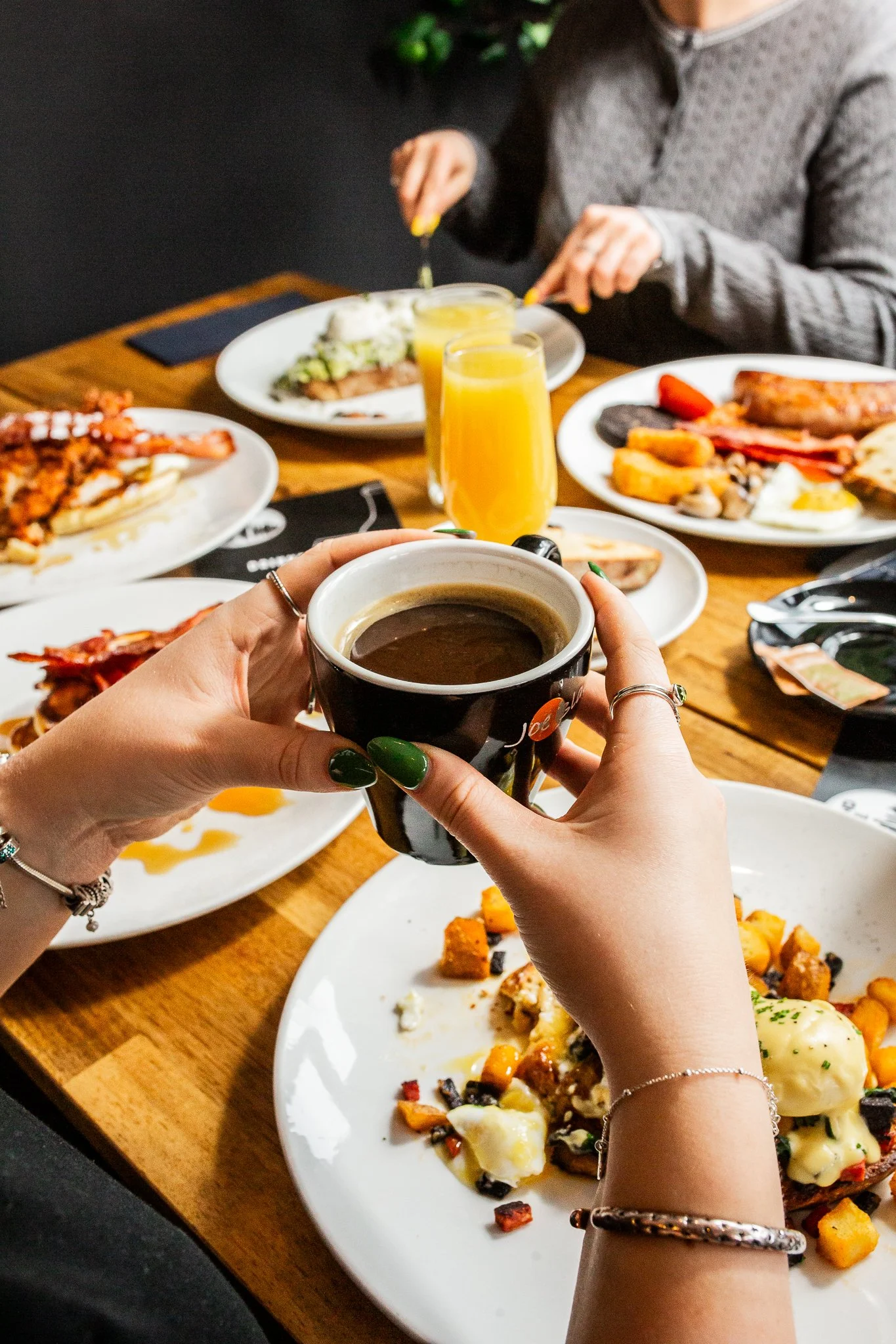 Person holding a cup of coffee over a table with assorted breakfast dishes, including eggs, bacon, potatoes, fruit, and glasses of orange juice, while others are dining in the background.