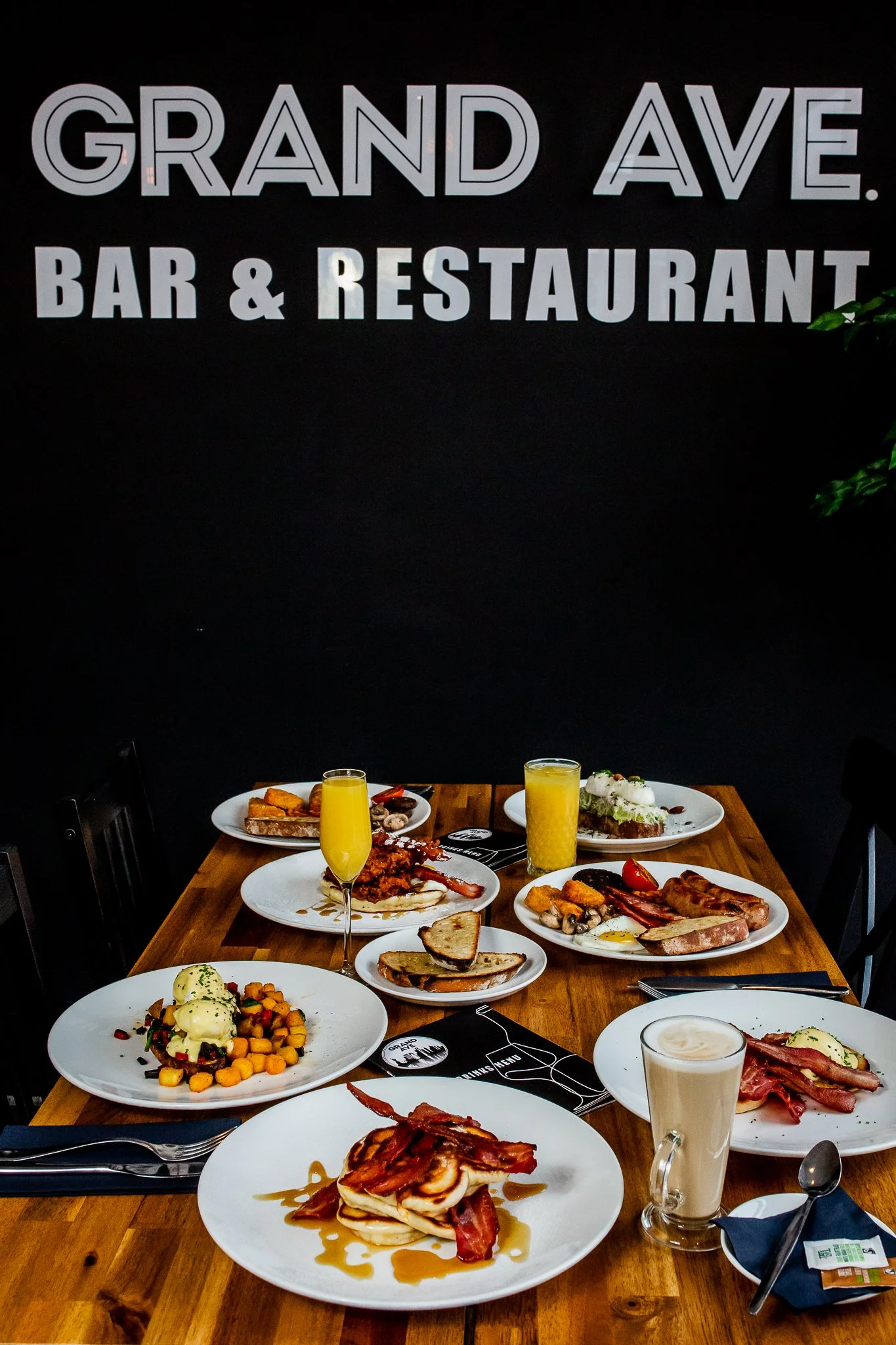 Table set with various breakfast dishes including eggs, bacon, toast, and drinks in a restaurant with a sign that reads 'Grand Ave. Bar & Restaurant' on the black wall behind.
