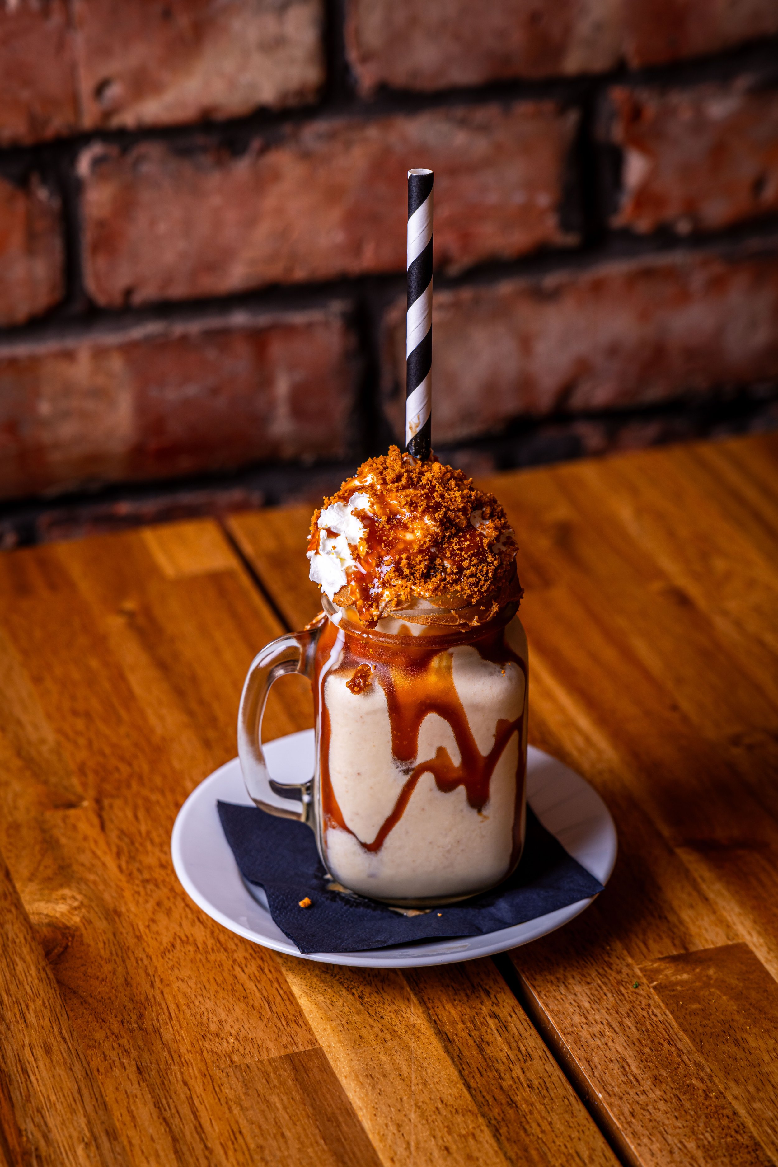 Frosted milkshake topped with whipped cream, caramel drizzle, and crumbled cookies in a glass jar with a striped straw, served on a white plate with a black napkin, on a wooden table against a brick wall background.