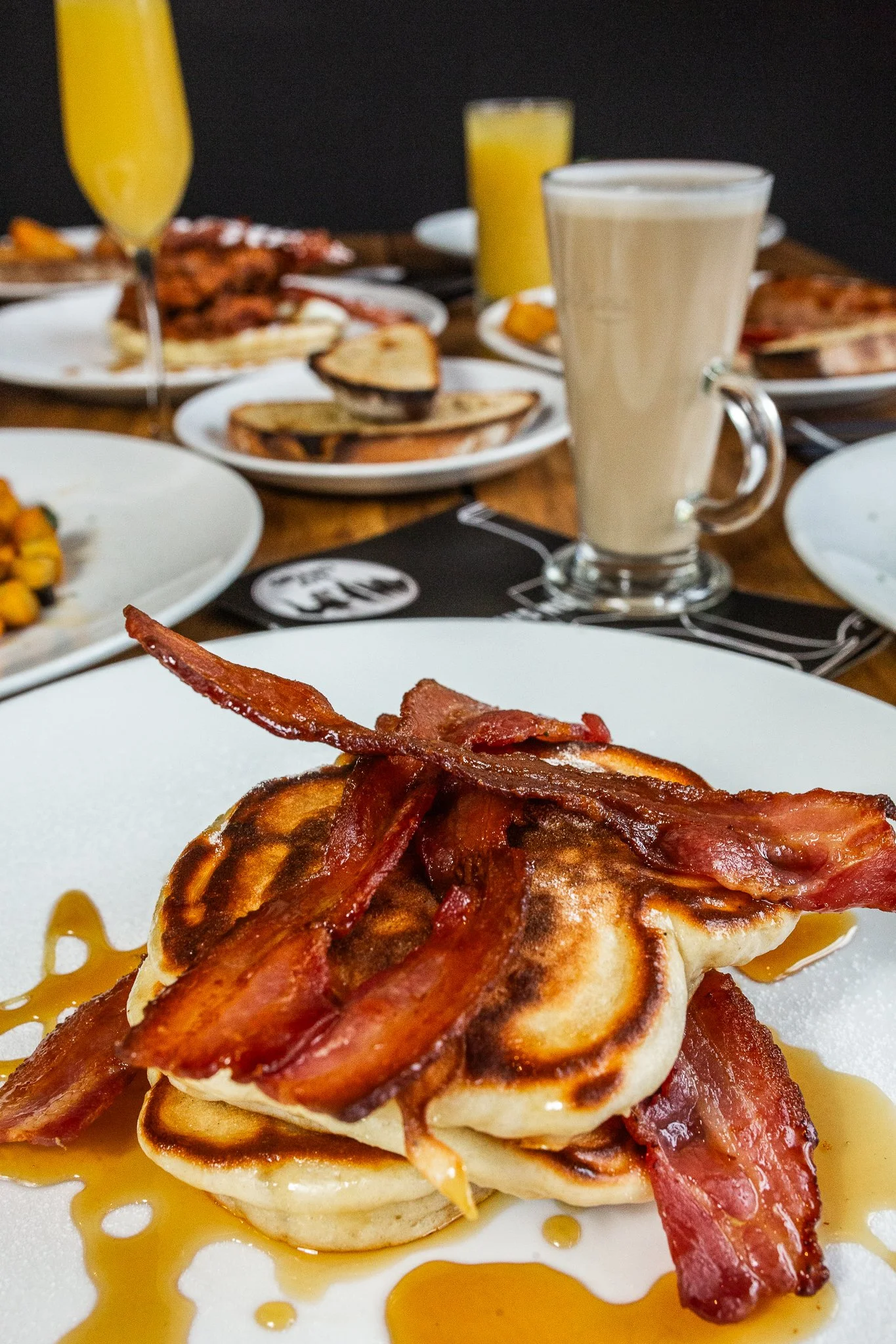 Close-up of a breakfast dish with pancakes topped with crispy bacon on a white plate, drizzled with syrup. In the background, a table has various breakfast foods, drinks, including a glass of orange juice, a latte, and other plates with food.