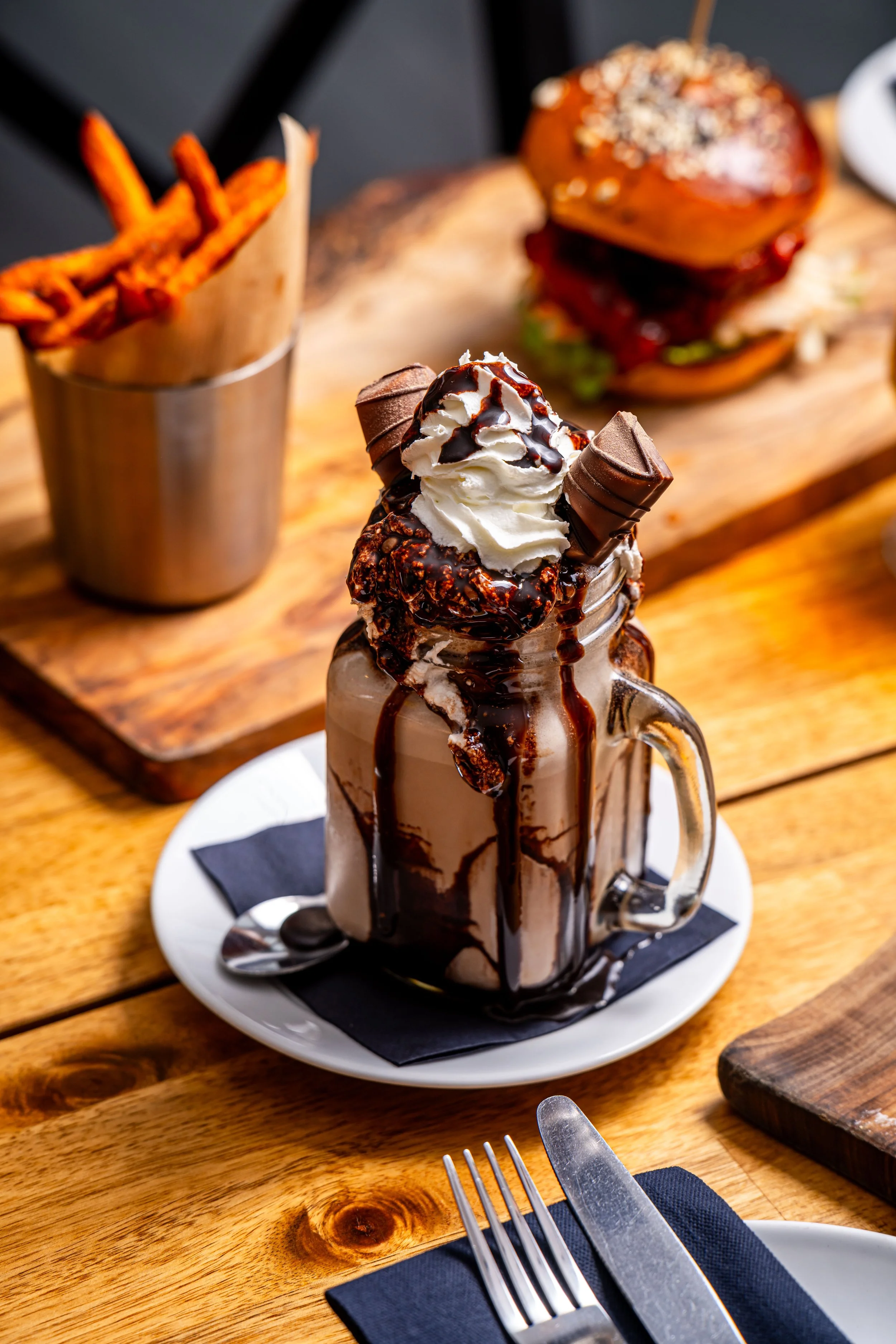 Chocolate milkshake topped with whipped cream, chocolate syrup, and two Rolos, served in a glass jar mug on a white plate with a black napkin and a spoon on a wooden table.