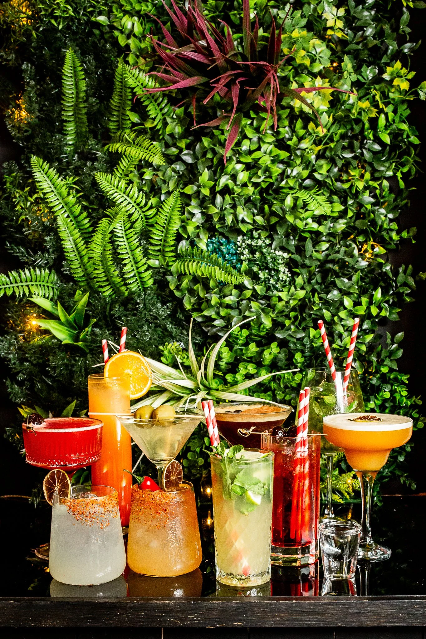 A colorful assortment of cocktails and drinks with straws, garnishes, and fruit slices on a black surface, with a lush green foliage background.