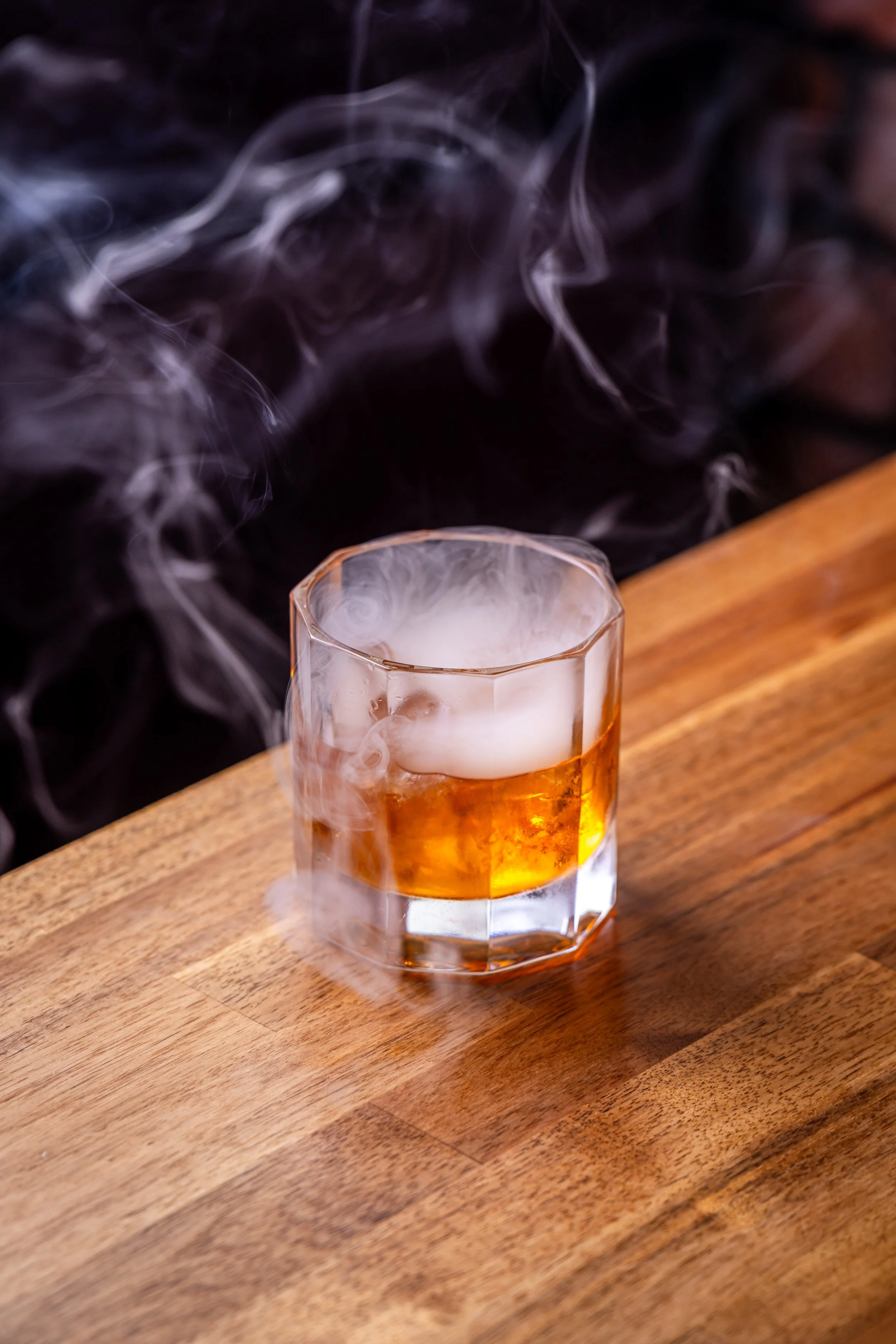 A glass of whiskey with smoke swirling around it on a wooden table.