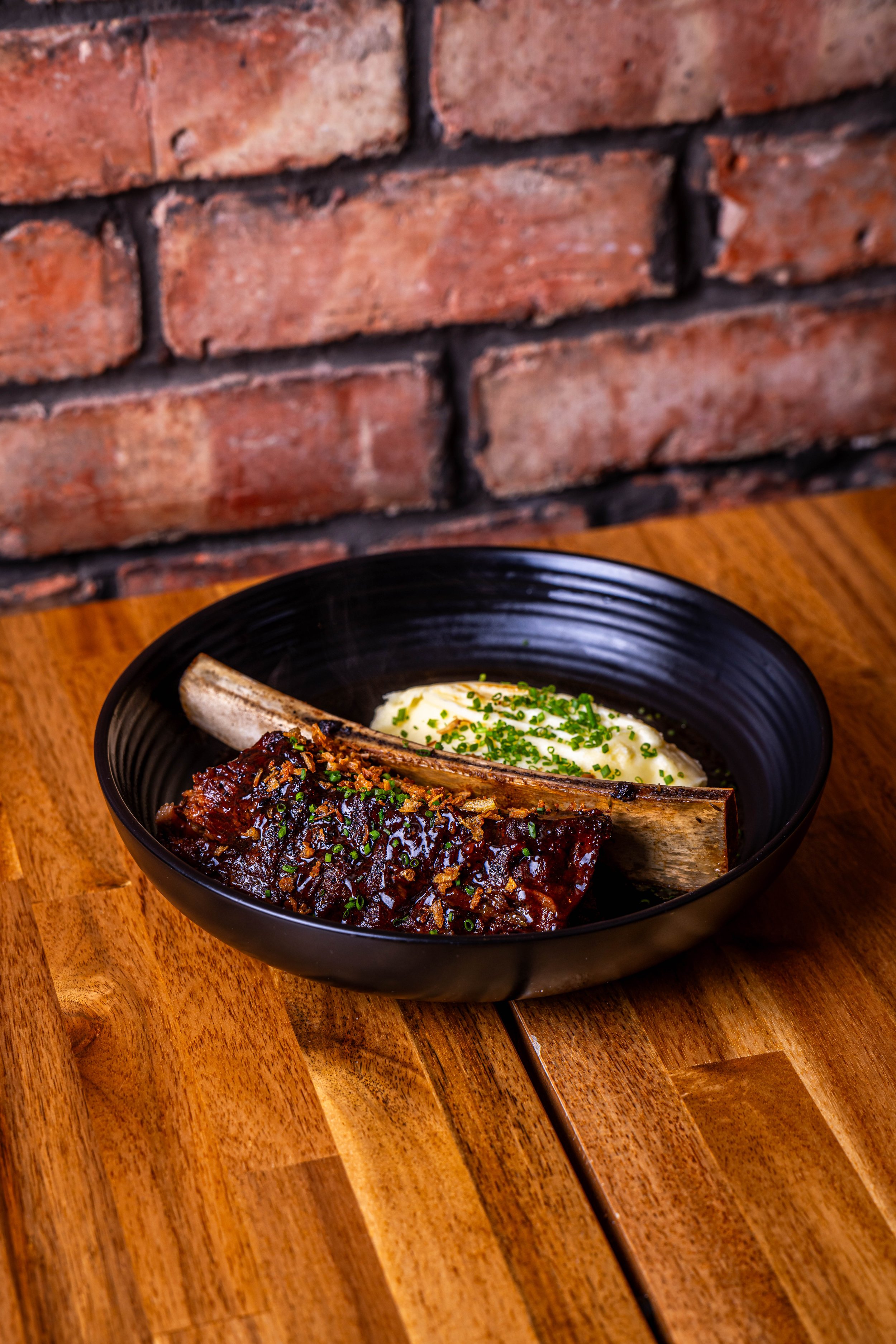 A black bowl containing a serving of braised short ribs with mashed potatoes garnished with chopped chives, on a wooden table against a brick wall background.