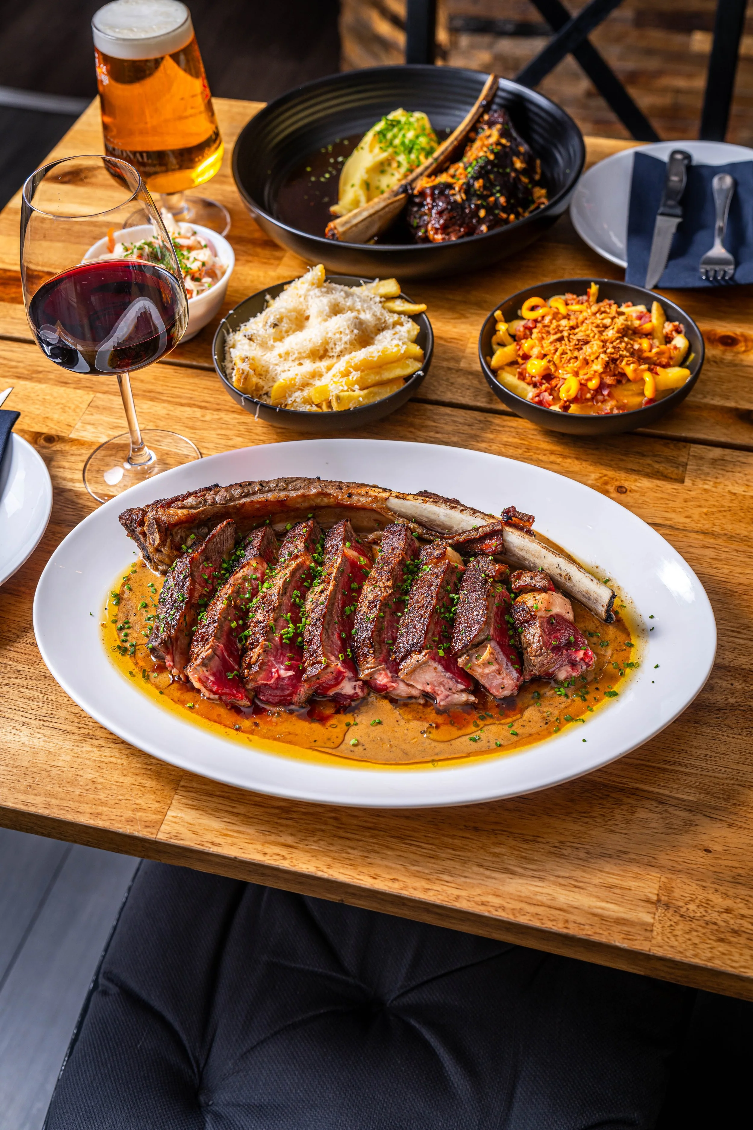 A dinner table with a large plate of sliced cooked steak with sauce, accompanied by a glass of red wine, a glass of beer, and various side dishes including macaroni and cheese, pasta with sauce, and a bowl of salad.