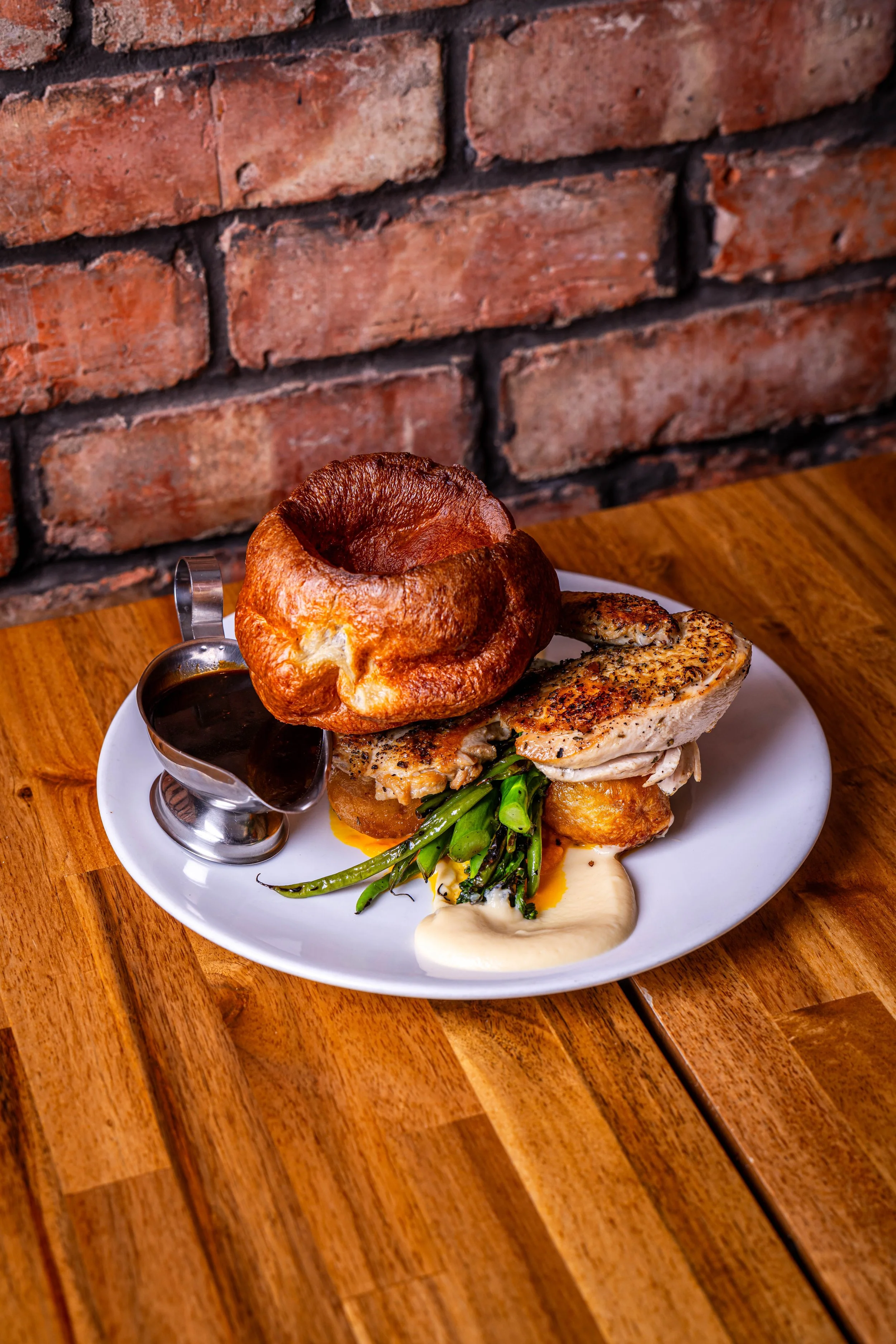 Plate of gourmet food with a Yorkshire pudding, grilled chicken breast, green beans, and creamy sauce, served with gravy on the side at a rustic restaurant.
