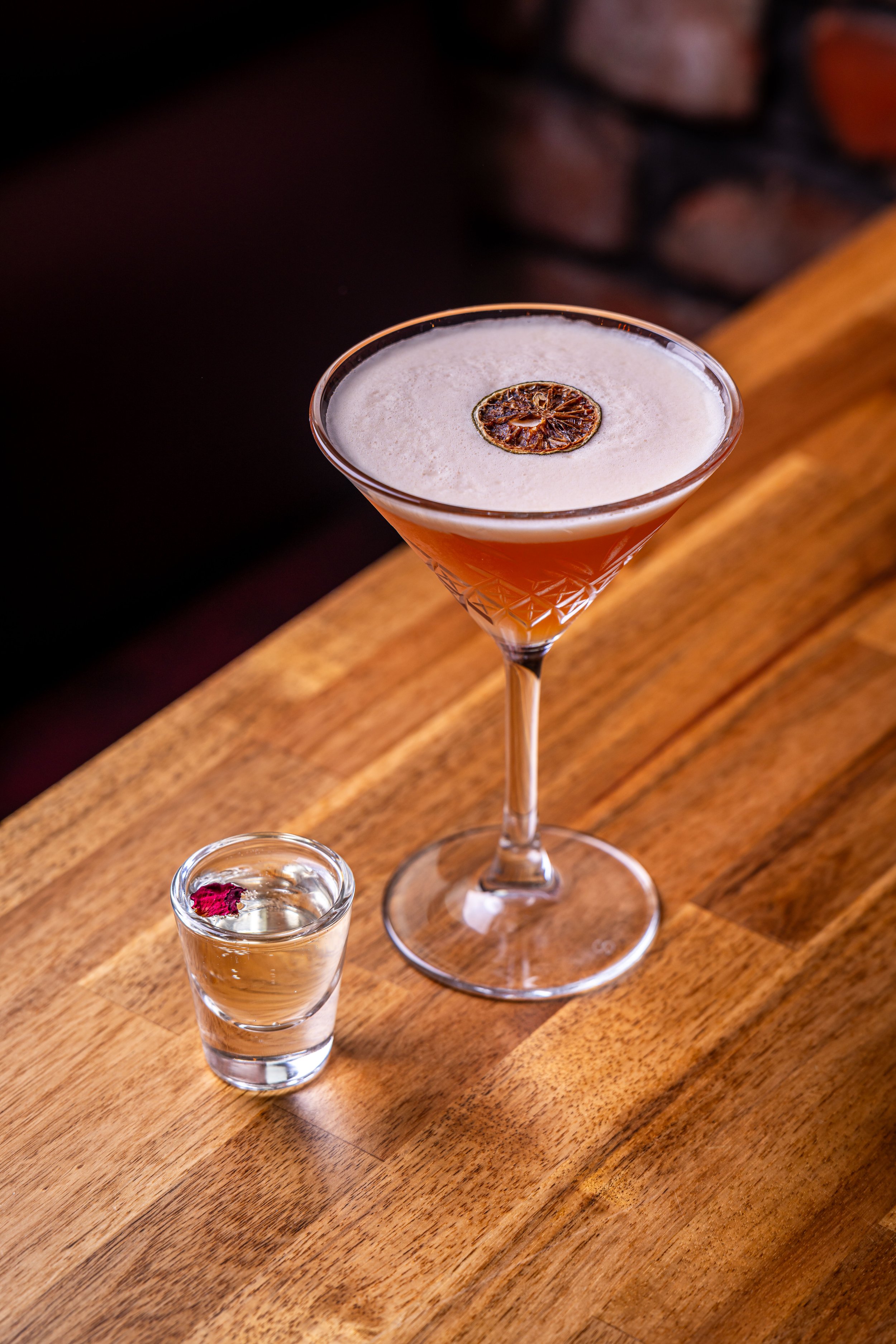 A cocktail in a martini glass with a dried citrus slice on top, placed on a wooden bar counter, with a small glass of clear liquid garnished with a red flower petal nearby.