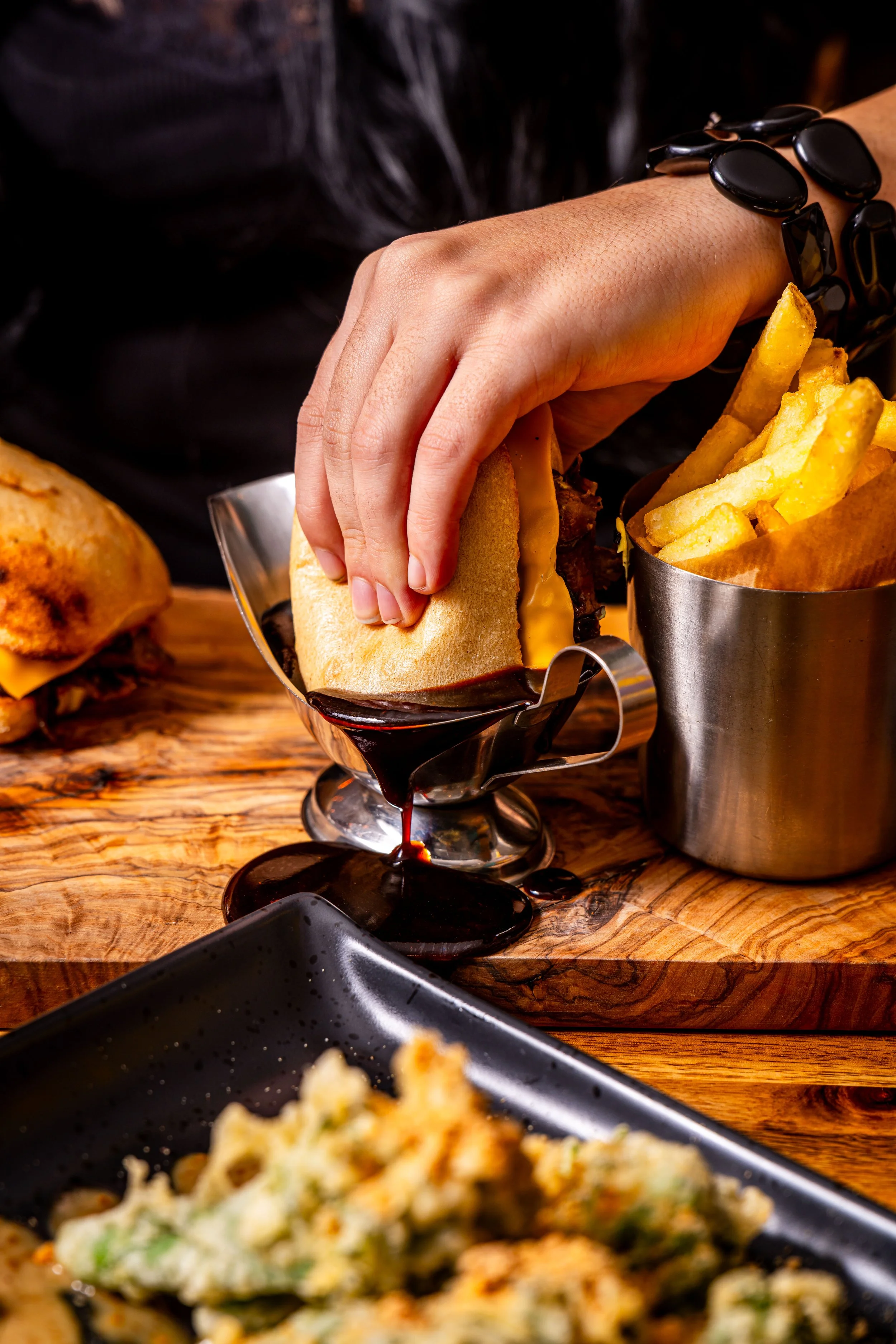 Hand holding a cheeseburger with cheese, along with a side of French fries in a metal container on a wooden table.