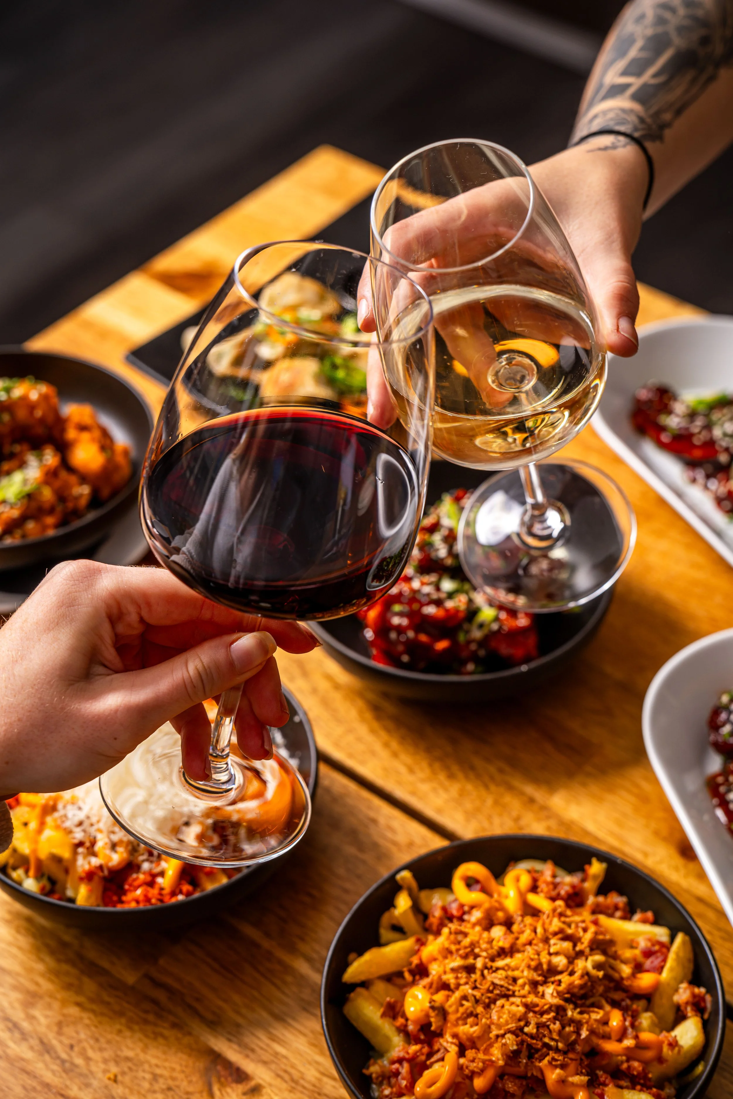 Two people clinking glasses of red and white wine over a table filled with bowls of food.