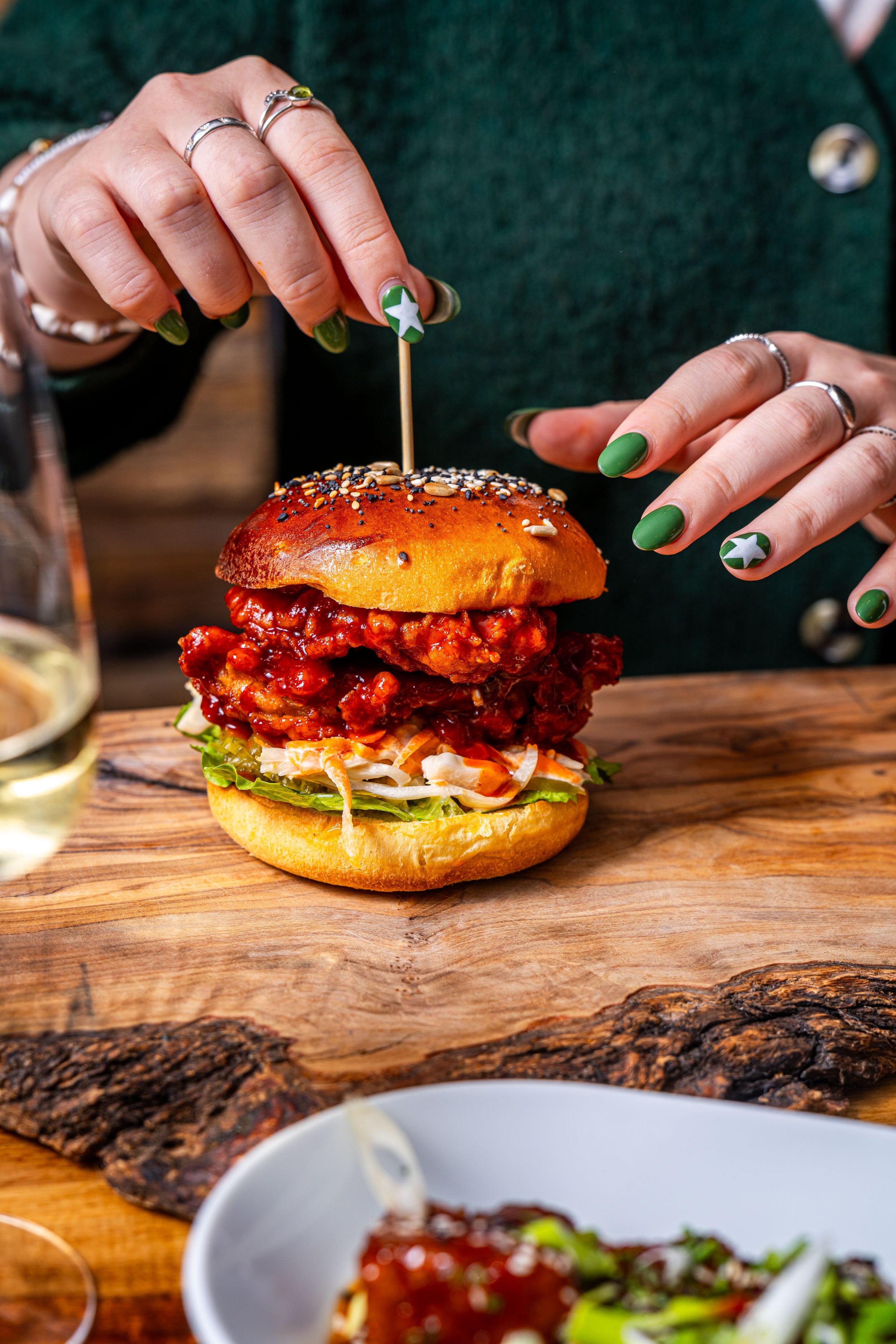 A person is placing a toothpick into a large, spicy fried chicken sandwich on a sesame seed bun, with lettuce and coleslaw, on a wooden table.