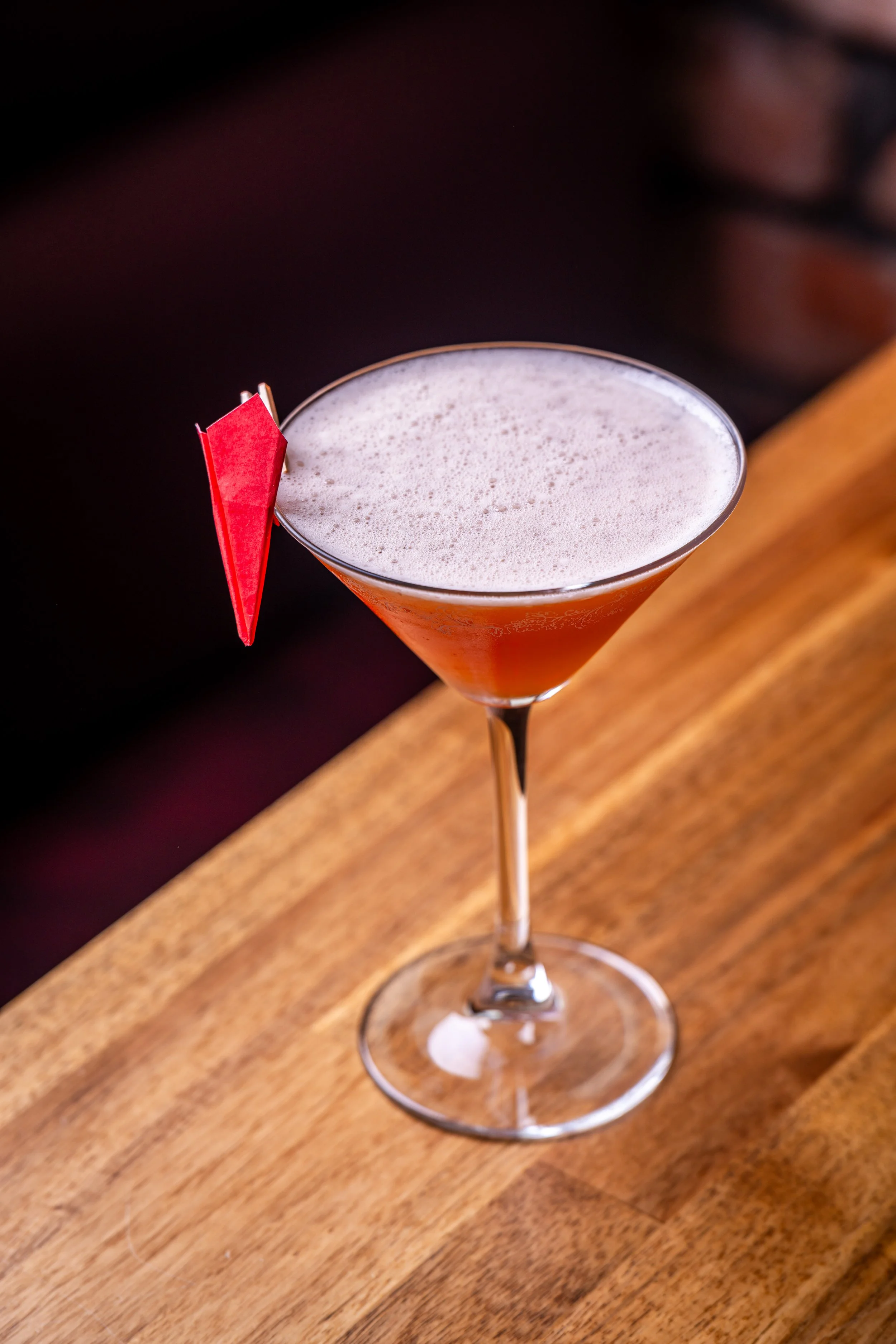 A cocktail in a martini glass with a foam top, garnished with a small red paper decoration on the rim, placed on a wooden bar counter.