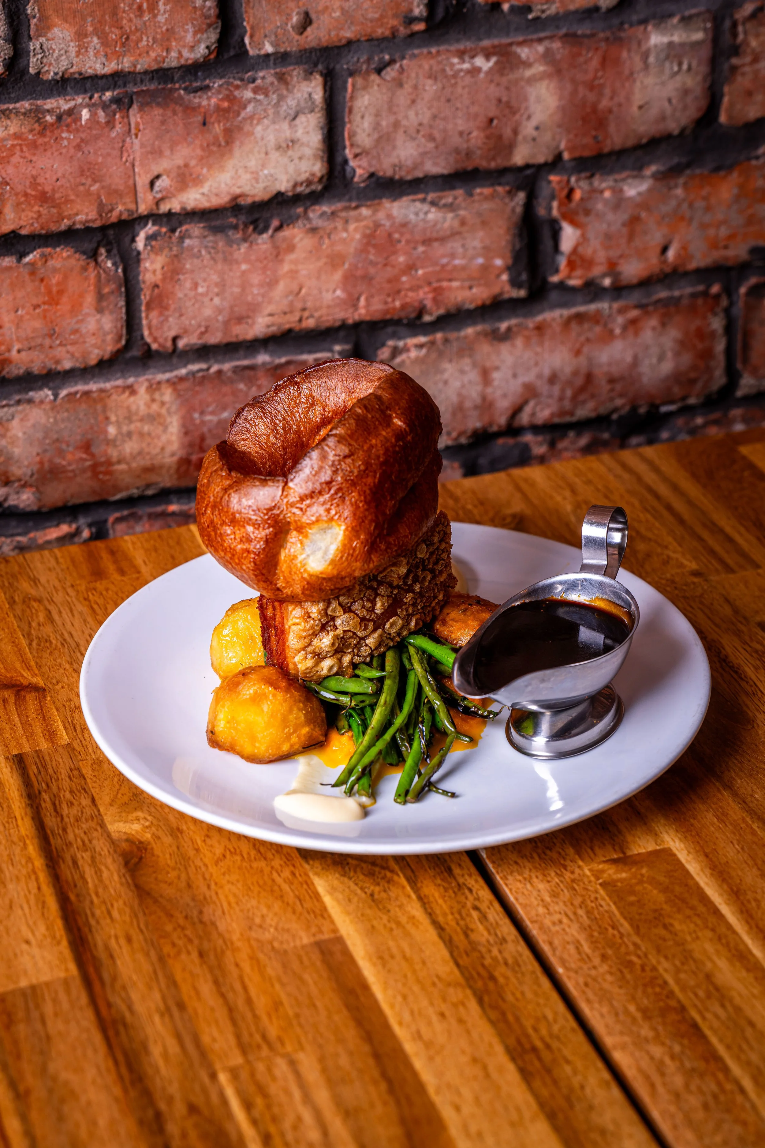 A plate of food with fried chicken, green beans, potatoes, a biscuit, and gravy on a white plate on a wooden table against a brick wall background.