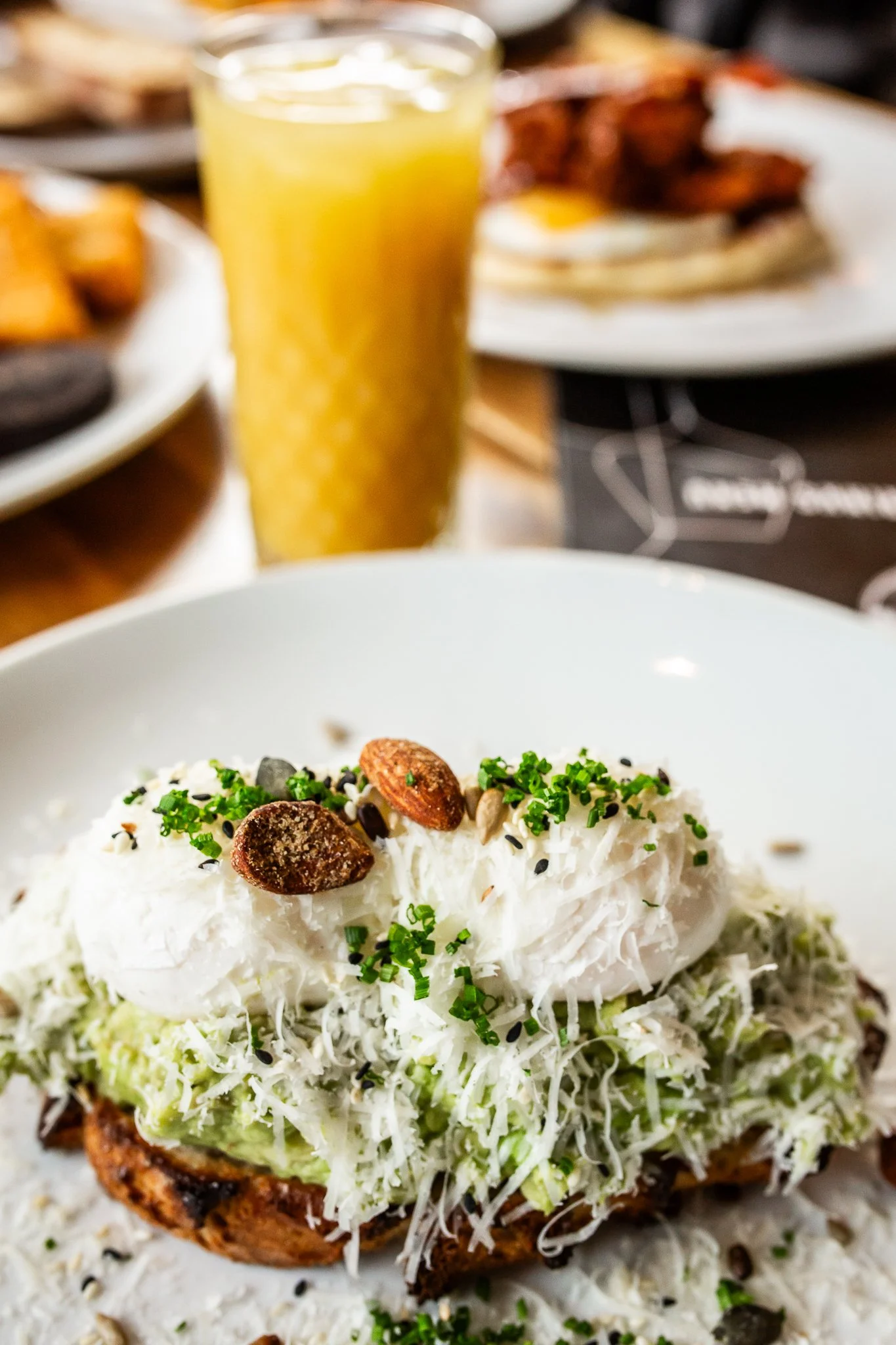 A close-up of an avocado toast topped with shredded cheese, chopped chives, and toasted almonds, served on a white plate. In the background, a glass of orange juice and blurry plates of food are visible.