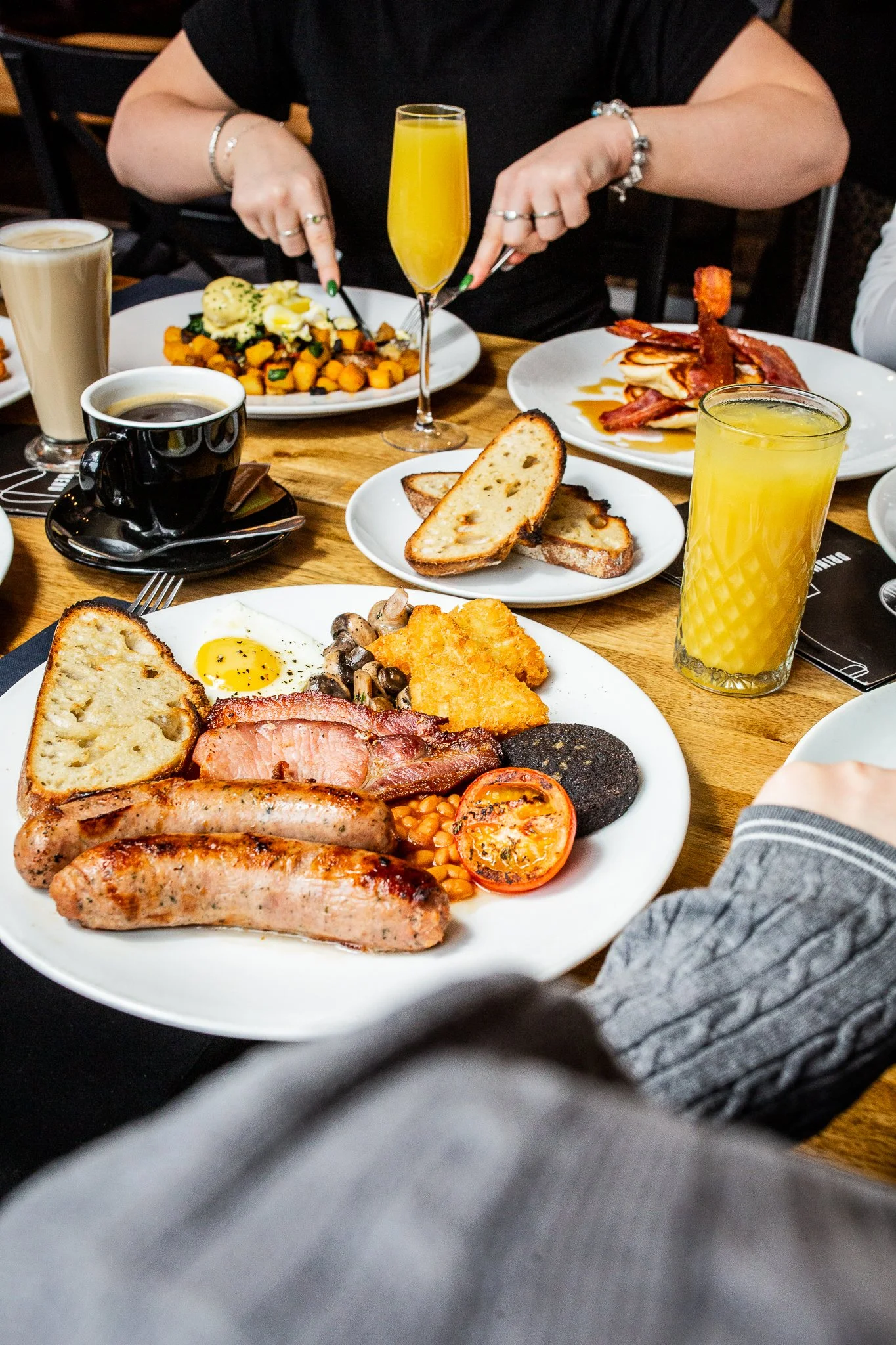 A breakfast table with plates of sausages, bacon, toast, eggs, grilled tomato, baked beans, mushrooms, and hash browns, along with glasses of orange juice and a cup of coffee.