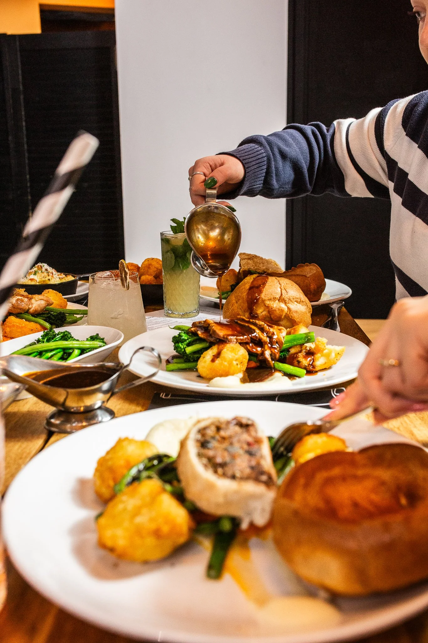 A table filled with various dishes, including a plate with a stuffed meat roll, greens, and potatoes, with a person pouring gravy over a plate of food and other plates of food and drinks in the background.