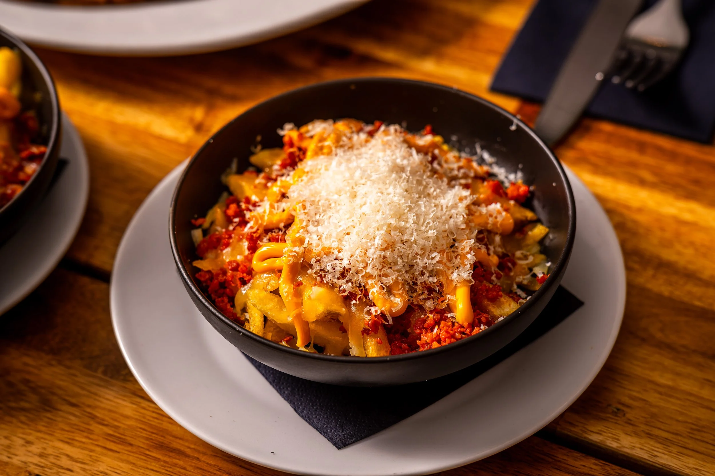 Bowl of seasoned fries topped with cheese, bacon bits, shredded cheese, and spicy sauce, placed on a white plate on a wooden table.