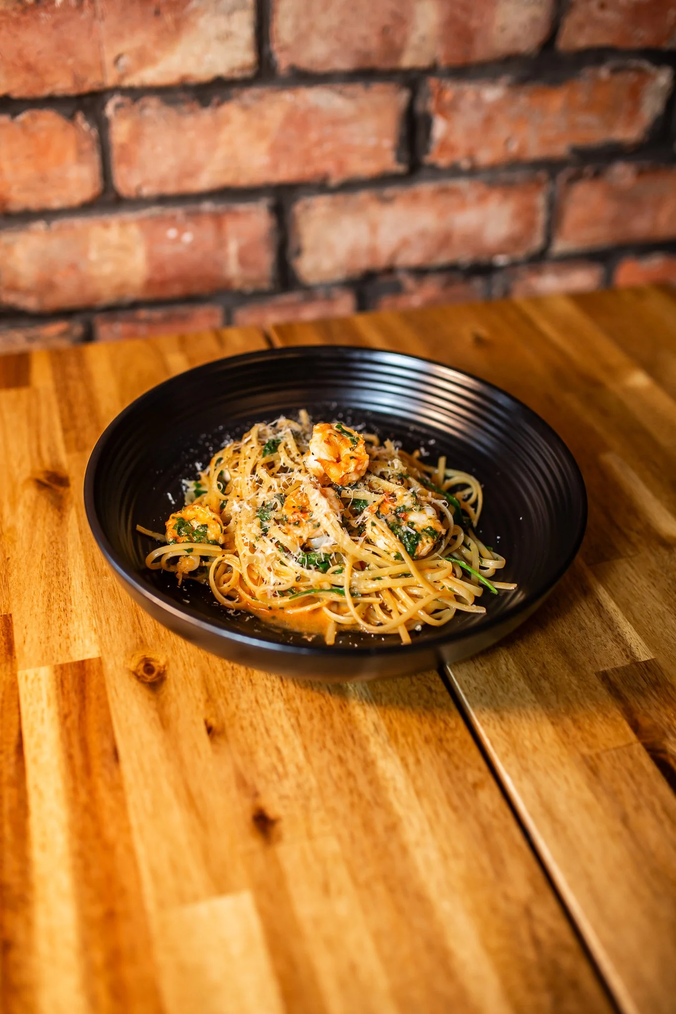 Black bowl of pasta with shrimp, grated cheese, and herbs on a wooden table against a brick wall.
