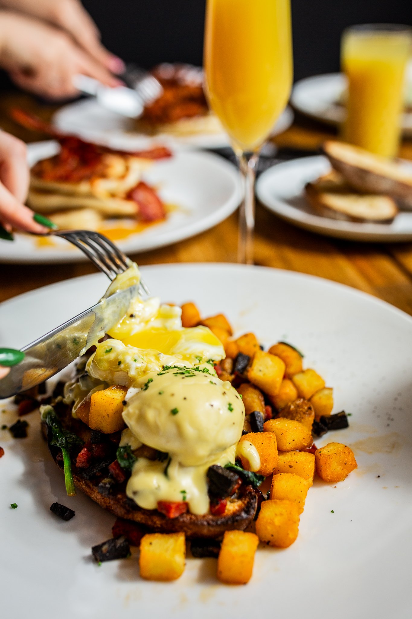 A plate of eggs Benedict with breakfast potatoes, with a glass of champagne and other plates of food in the background.
