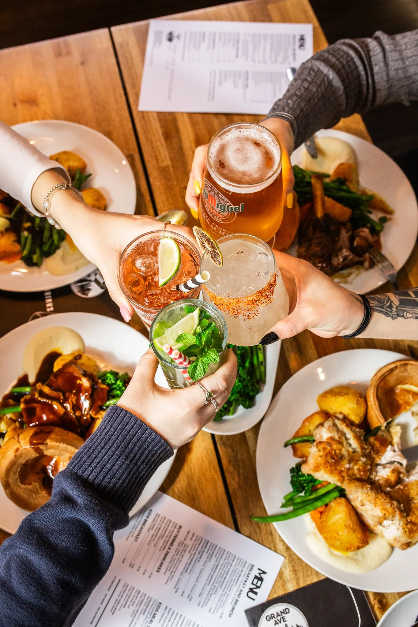 Four people clinking glasses of cocktails with food on the table, including plates of roasted chicken, potatoes, greens, and bread, in a restaurant setting.
