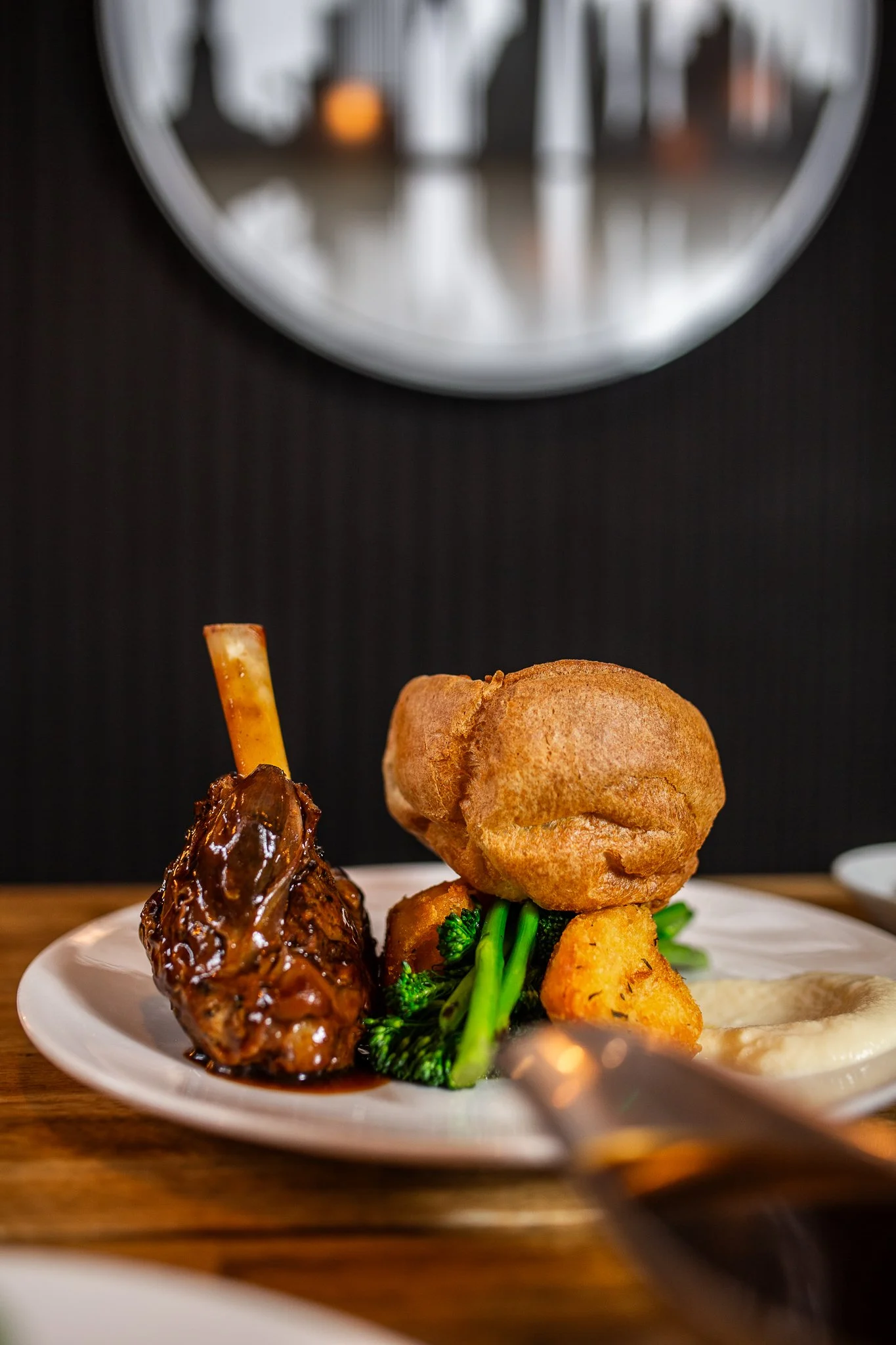 A plate of food featuring roasted chicken with barbecue sauce, broccoli, potato wedges, a Yorkshire pudding, and mashed potatoes, with a mirror reflection of the setting in the background.