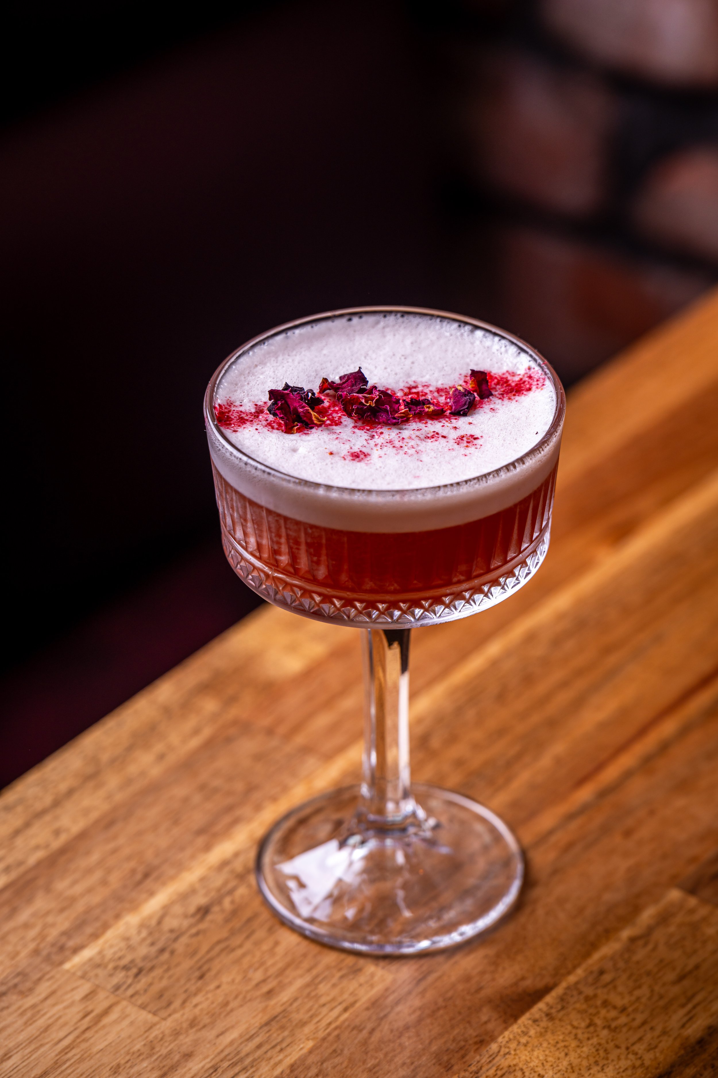 A cocktail served in a vintage glass with a textured pattern and a long stem, garnished with dried rose petals on top, placed on a wooden surface.