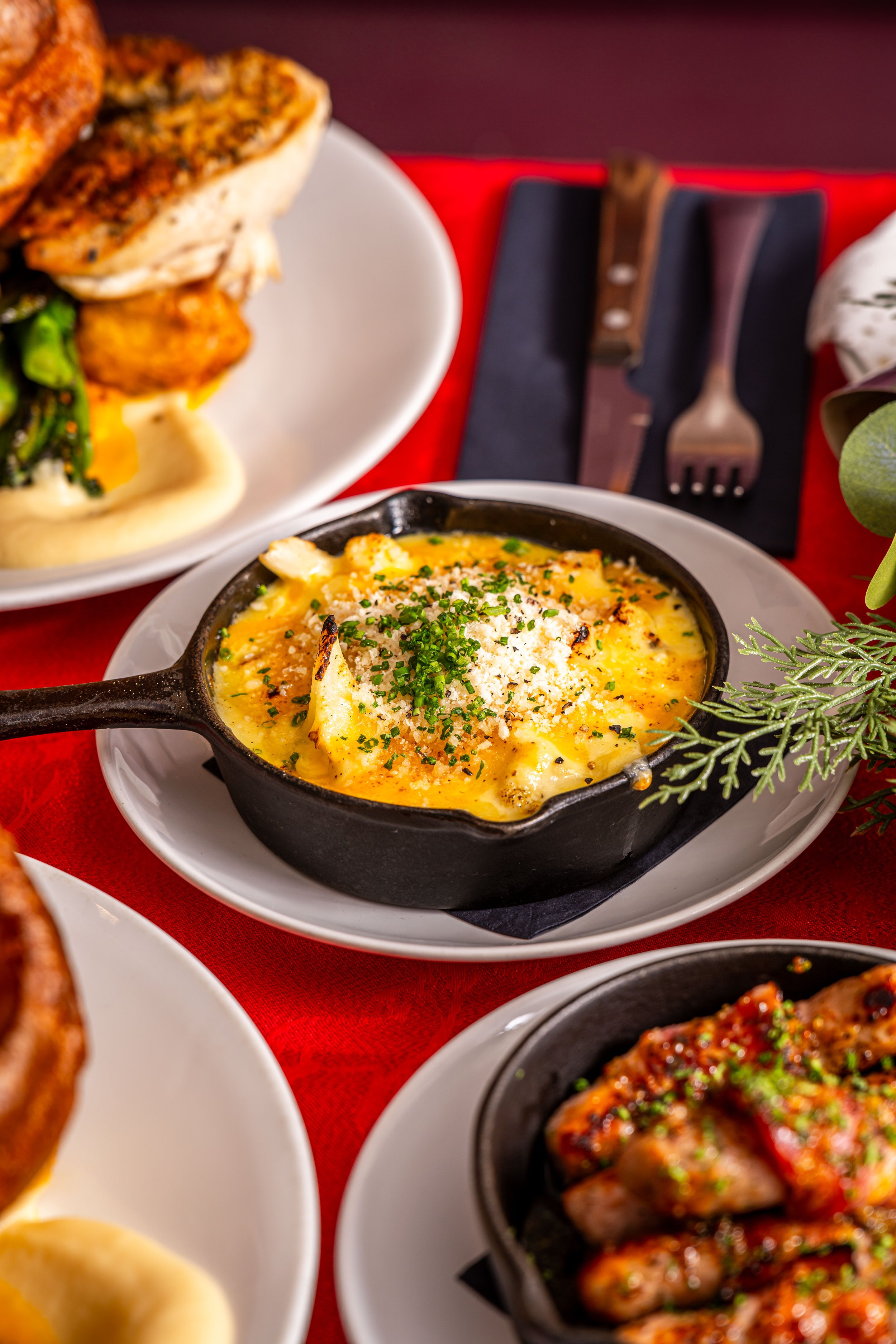 A serving of baked macaroni and cheese in a black cast iron skillet, topped with chopped chives and grated cheese, on a white plate. Part of other dishes, like grilled meat, are visible around it on a red tablecloth.