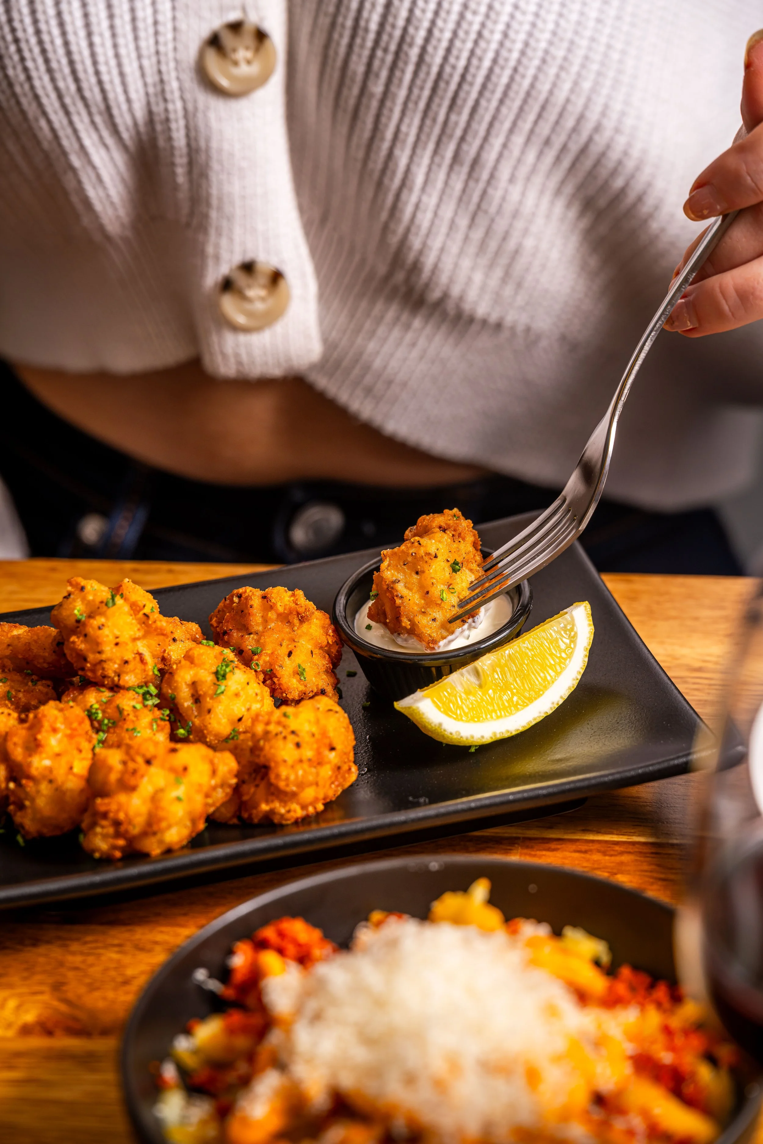 Fried cauliflower bites on a black plate with a lemon wedge and dipping sauce, person using fork to pick up a piece, wood table surface.