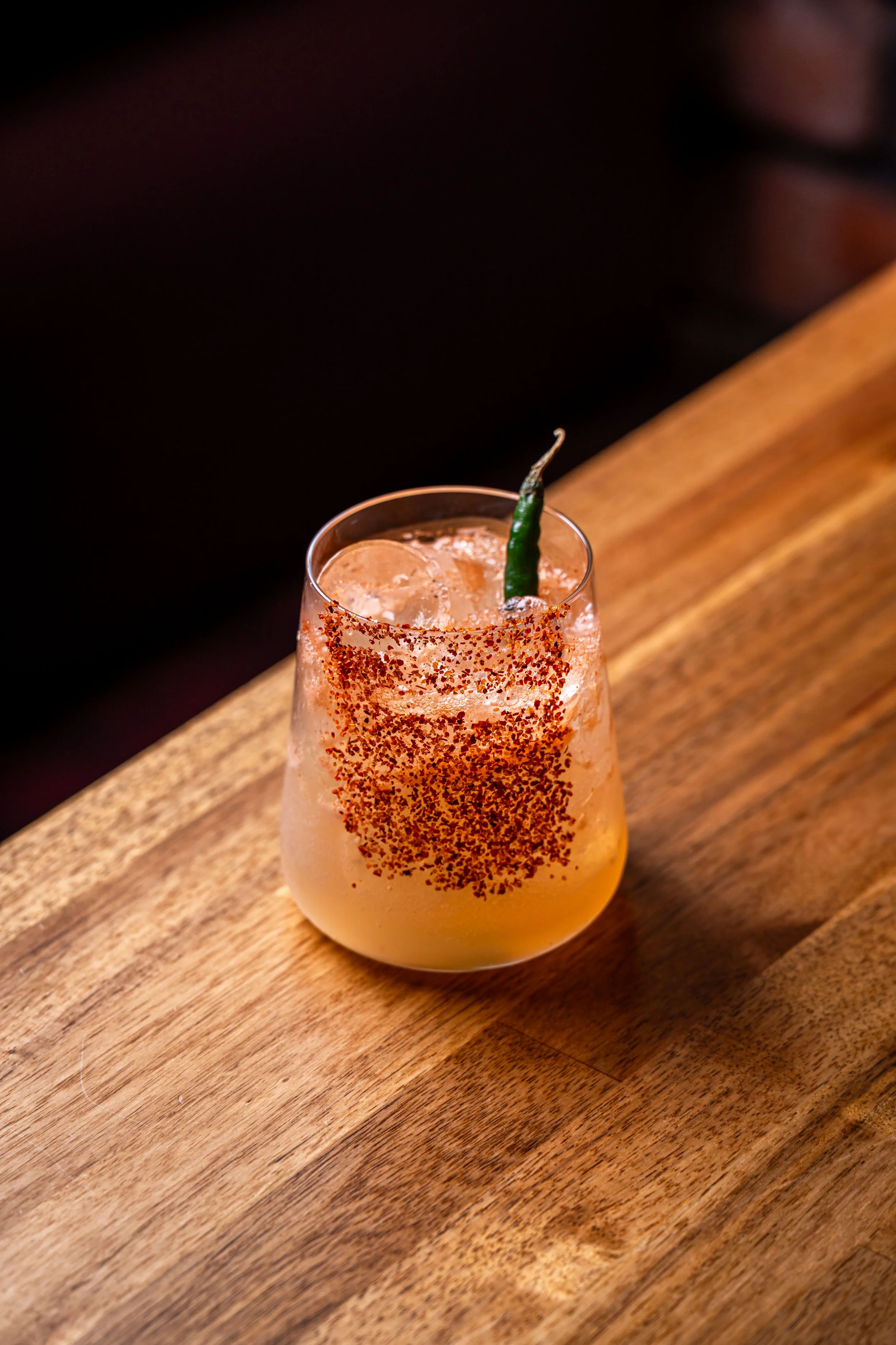Cocktail glass with ice, garnished with a green chili pepper and red chili powder on the rim, placed on a wooden surface.