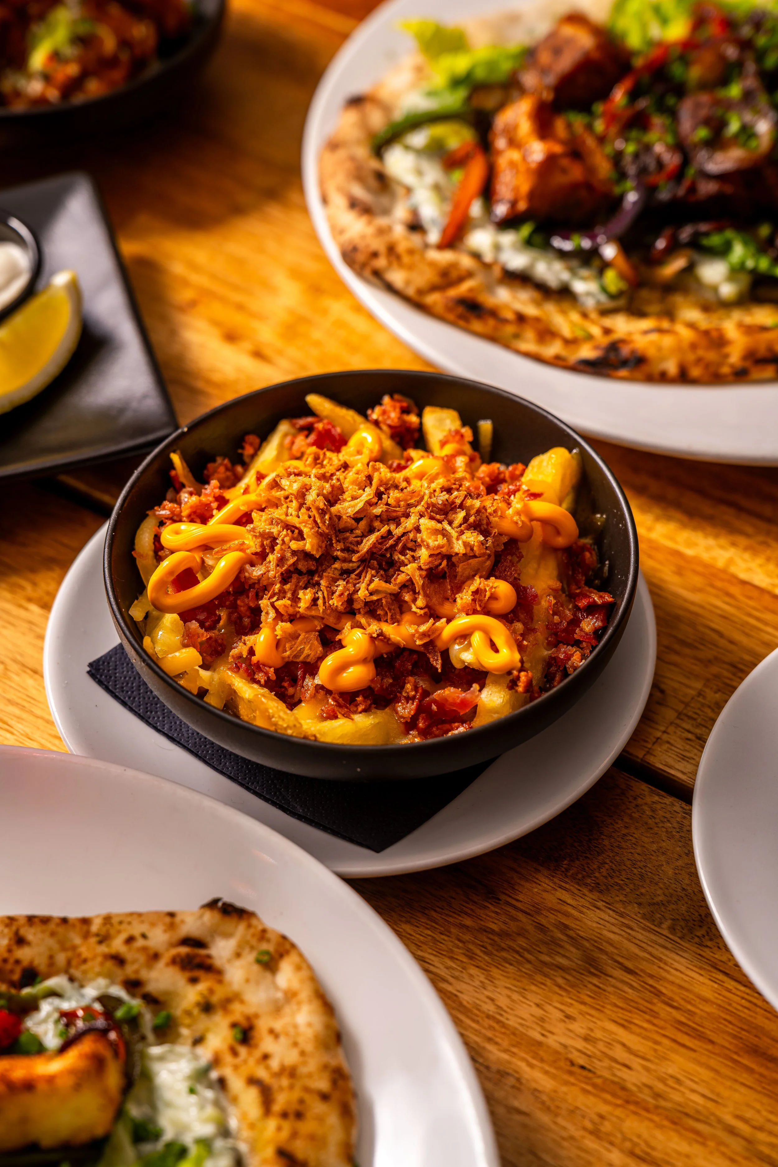 A bowl of loaded fries with cheese, bacon, and shredded meat on top, served on a black plate on a white saucer. In the background, there are plates of pizza and a dish with grilled meat and vegetables.