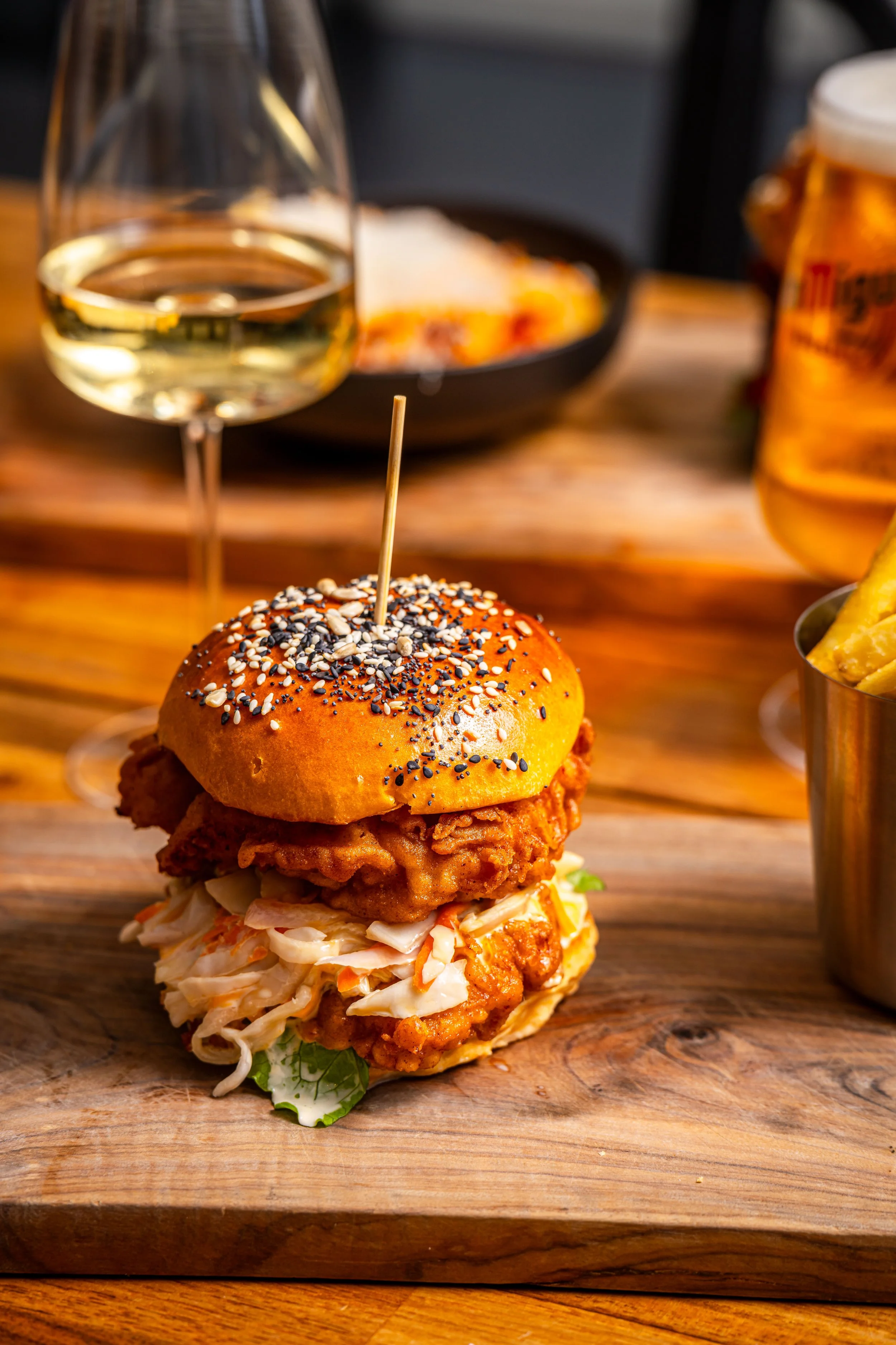 Fried chicken sandwich with lettuce and coleslaw in a sesame seed bun, served on a wooden plank, with a glass of white wine and a beer in the background.