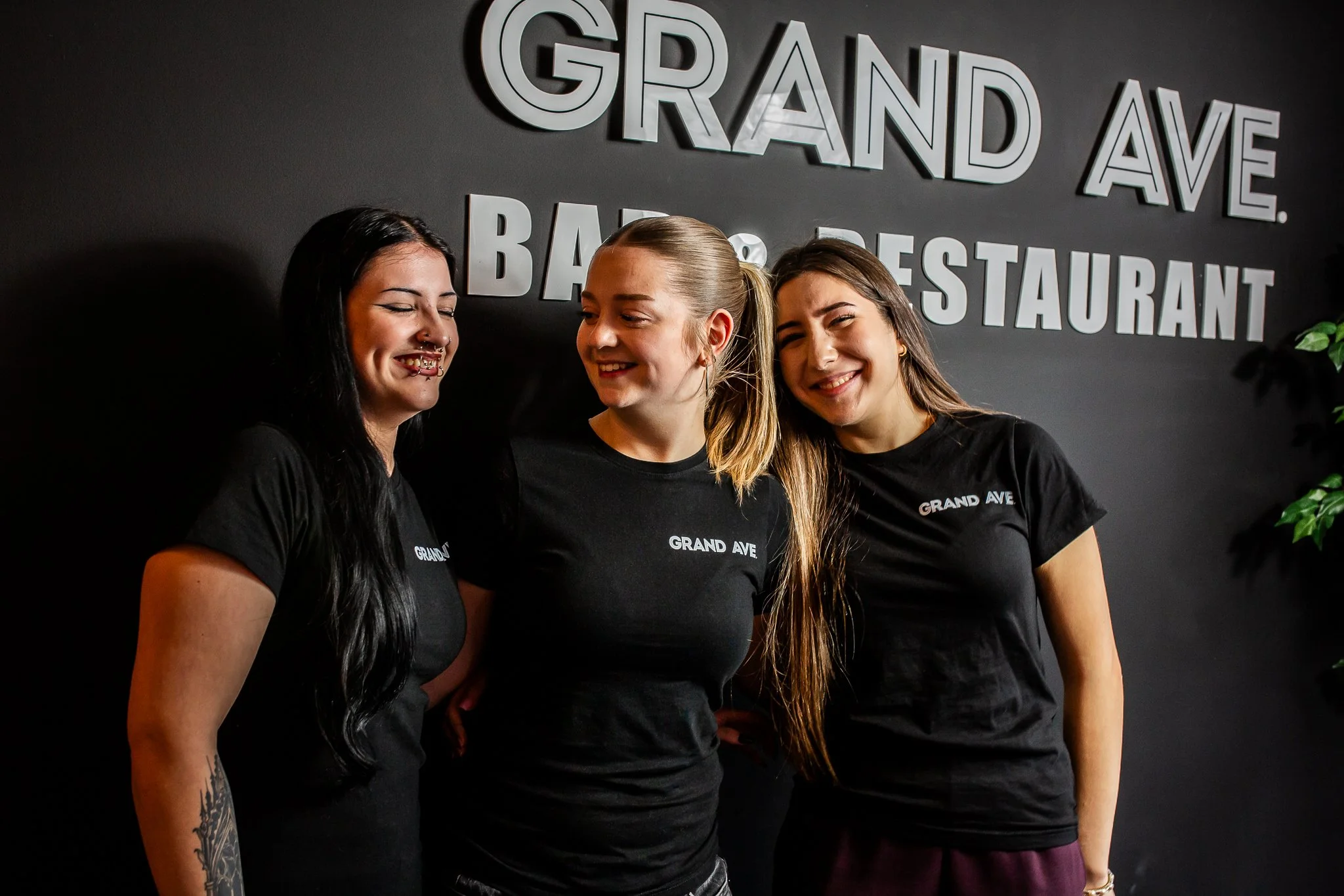 Three smiling women in black T-shirts standing close together in front of a dark wall with silver 3D letters that say 'Grand Ave' and 'Bar & Restaurant'.
