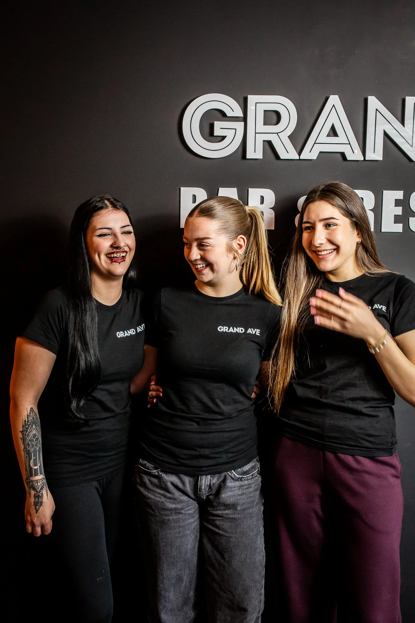 Three women wearing black "Grand Ave" t-shirts, standing together in front of a dark wall with large white letters saying "GRAND" and "BAR STRESS," smiling and laughing.