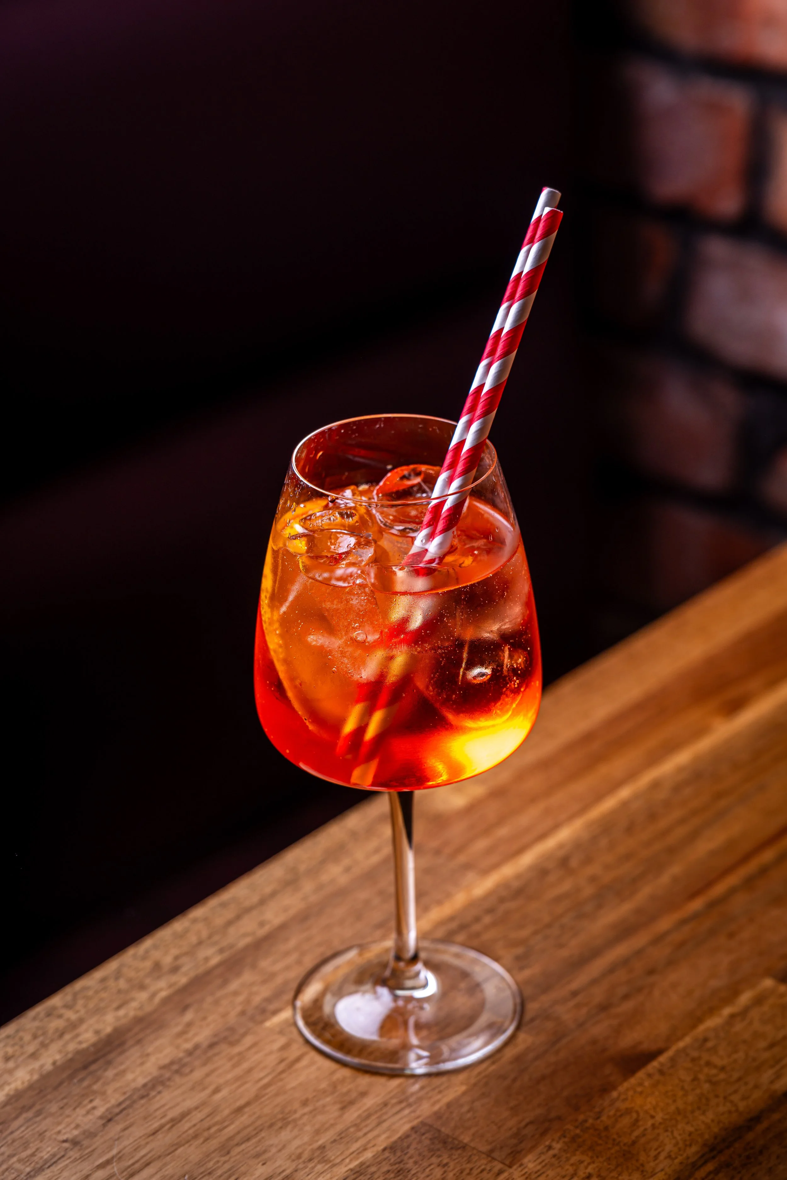 A glass of orange-colored cocktail with ice cubes and a red and white striped straw on a wooden surface.