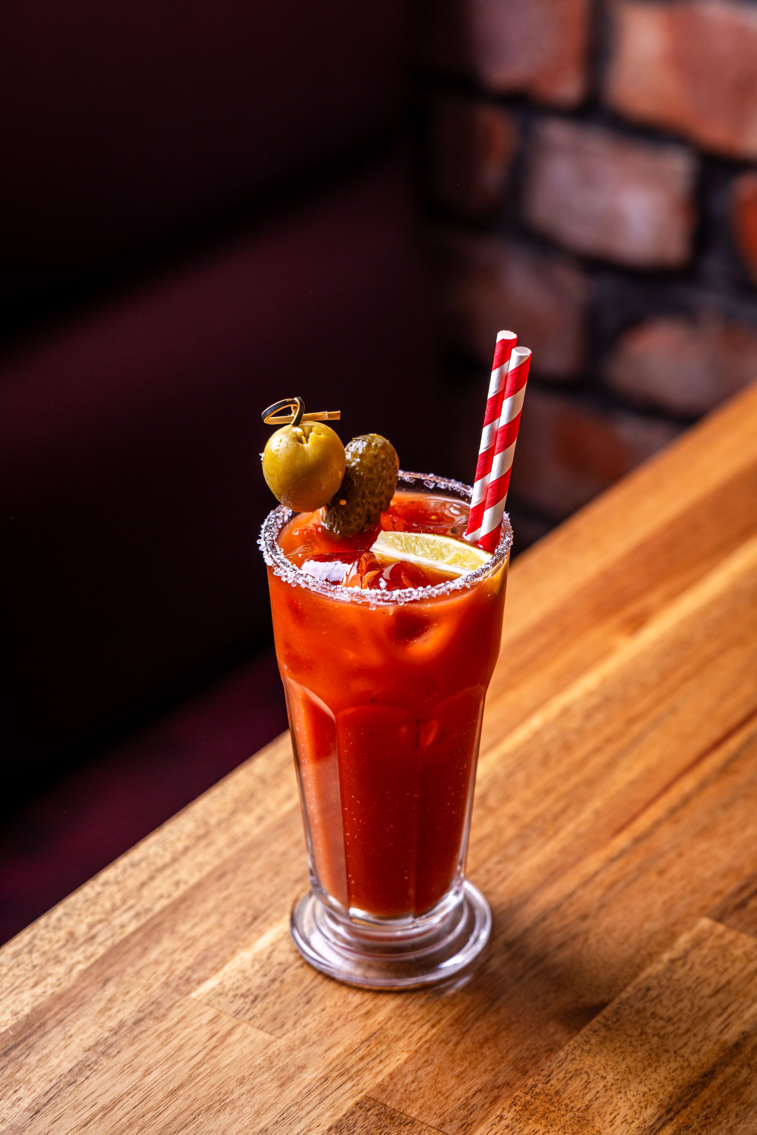 A tall glass of Bloody Mary garnished with an olive, a pickle, a lemon wedge, and two striped red and white straws on a wooden surface.