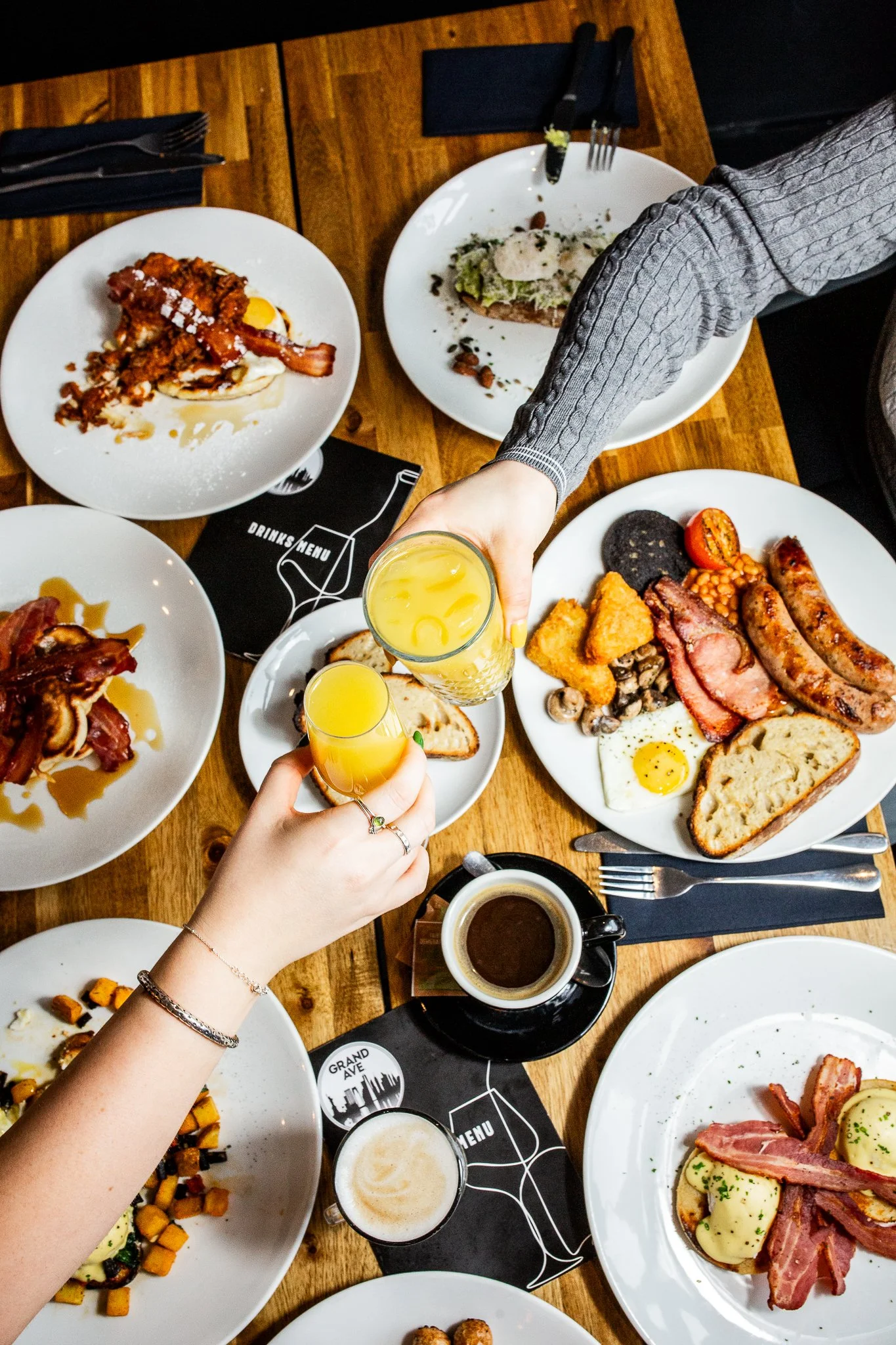 A top-down view of a wooden table with various plates of breakfast food, drinks, and a person's hands holding glasses for a toast.