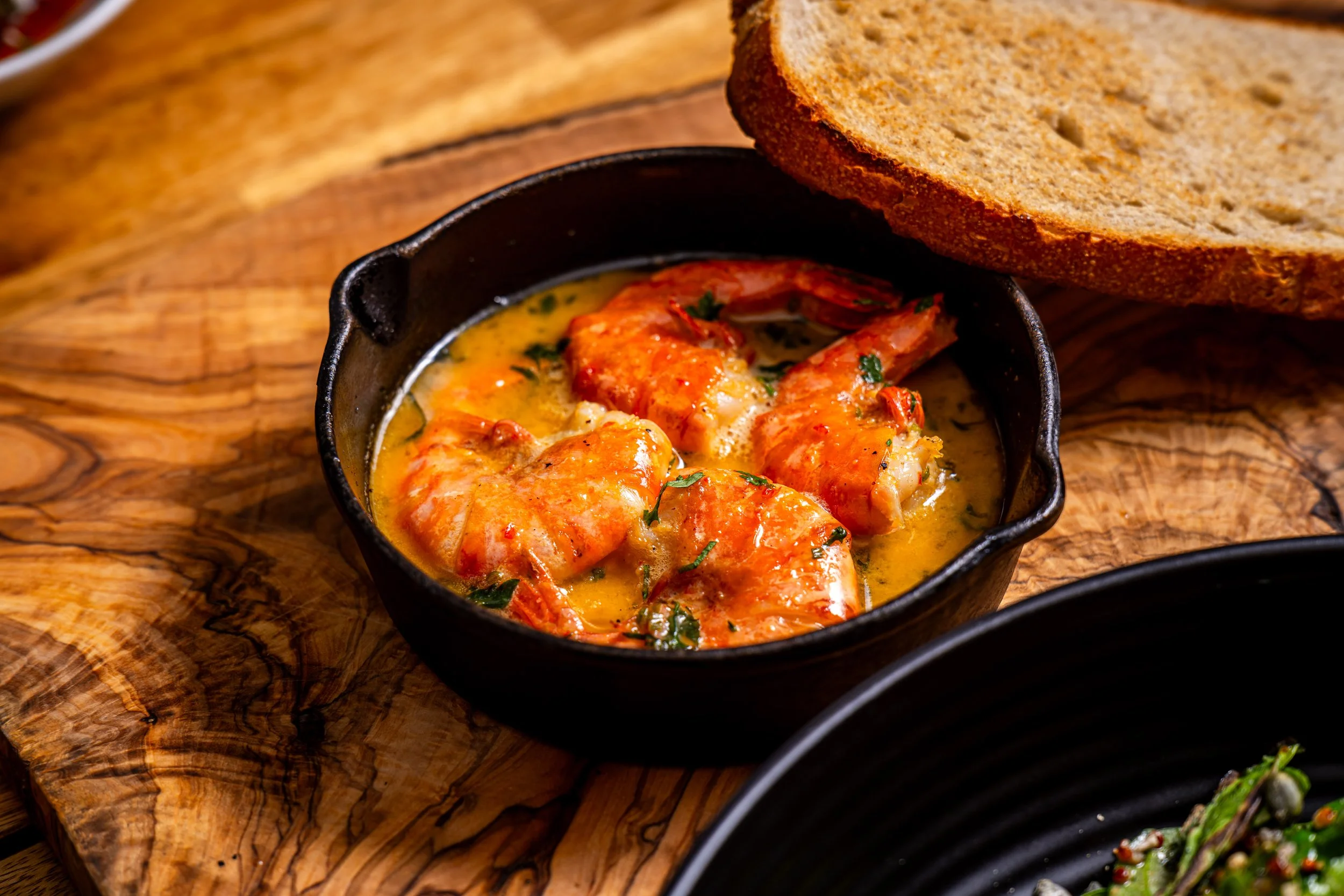 A black bowl filled with cooked shrimp in a creamy buttery sauce, garnished with chopped herbs, on a wooden surface with slices of bread nearby.