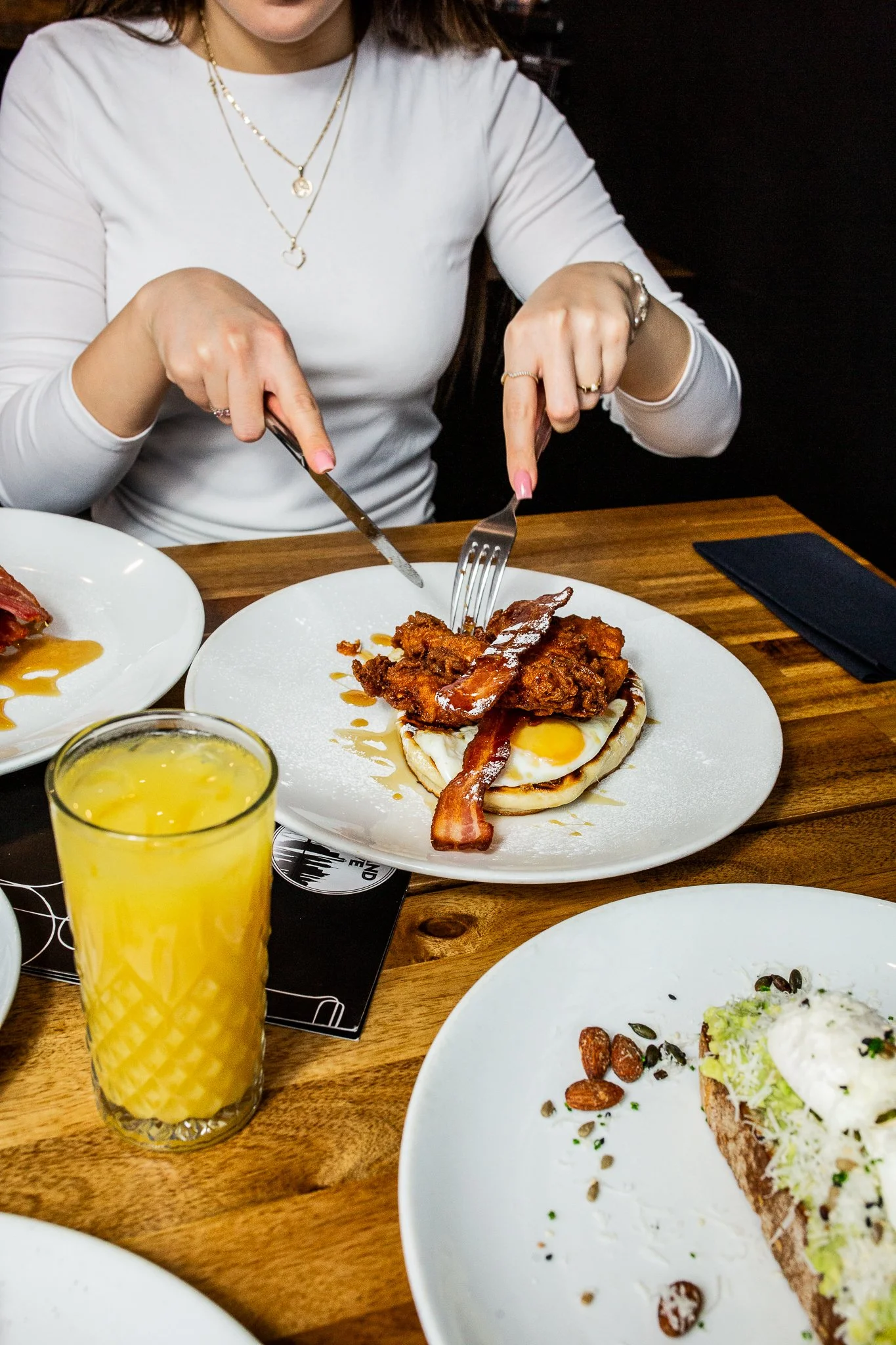 A woman in a white long-sleeve shirt is cutting into a breakfast dish that includes a fried egg, crispy bacon, and fried chicken on a white plate. The table also has a glass of orange juice and another plate with toast topped with avocado and cheese.