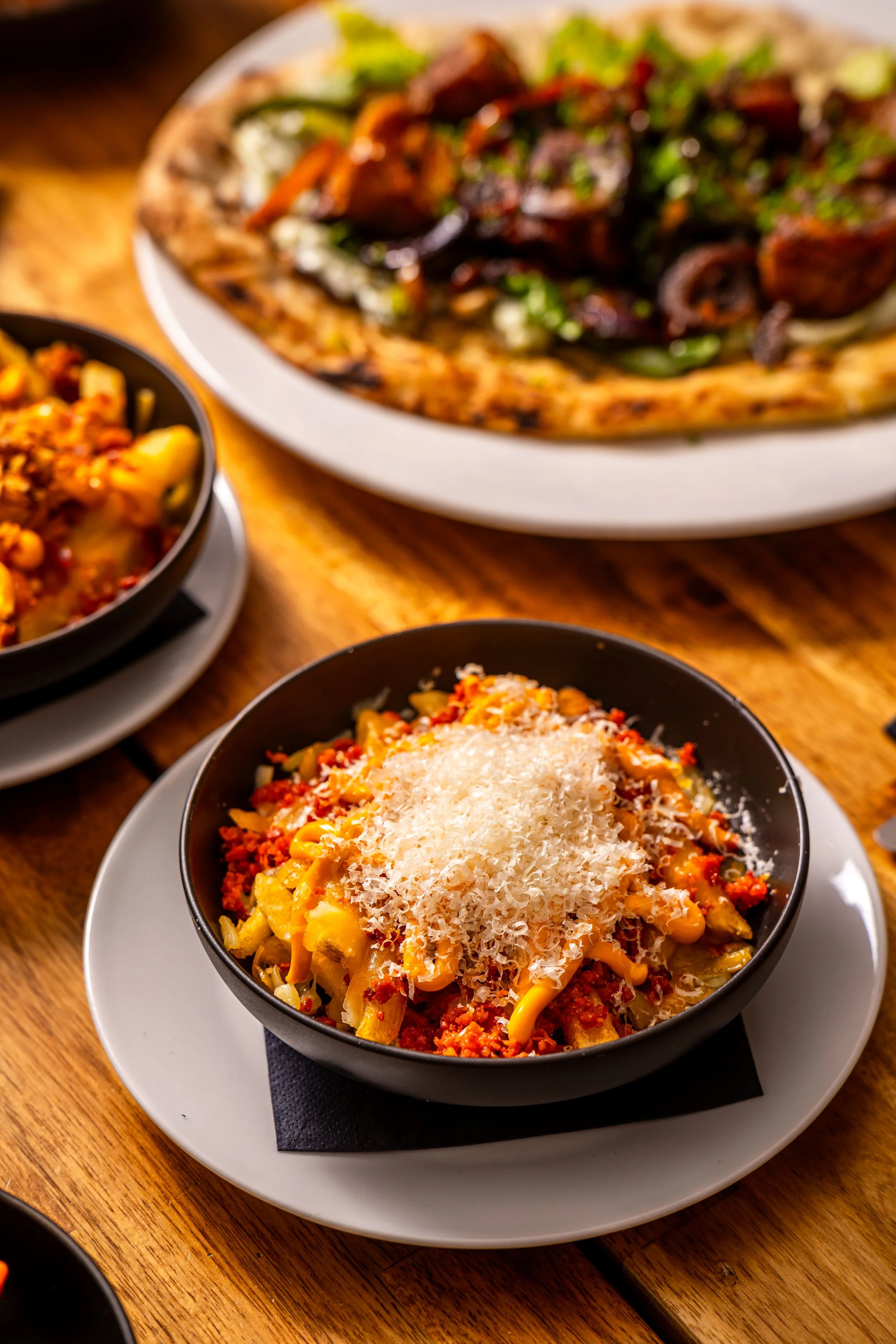 A bowl of macaroni and cheese topped with shredded cheese on a white plate, with a pizza and another dish in the background on a wooden table.
