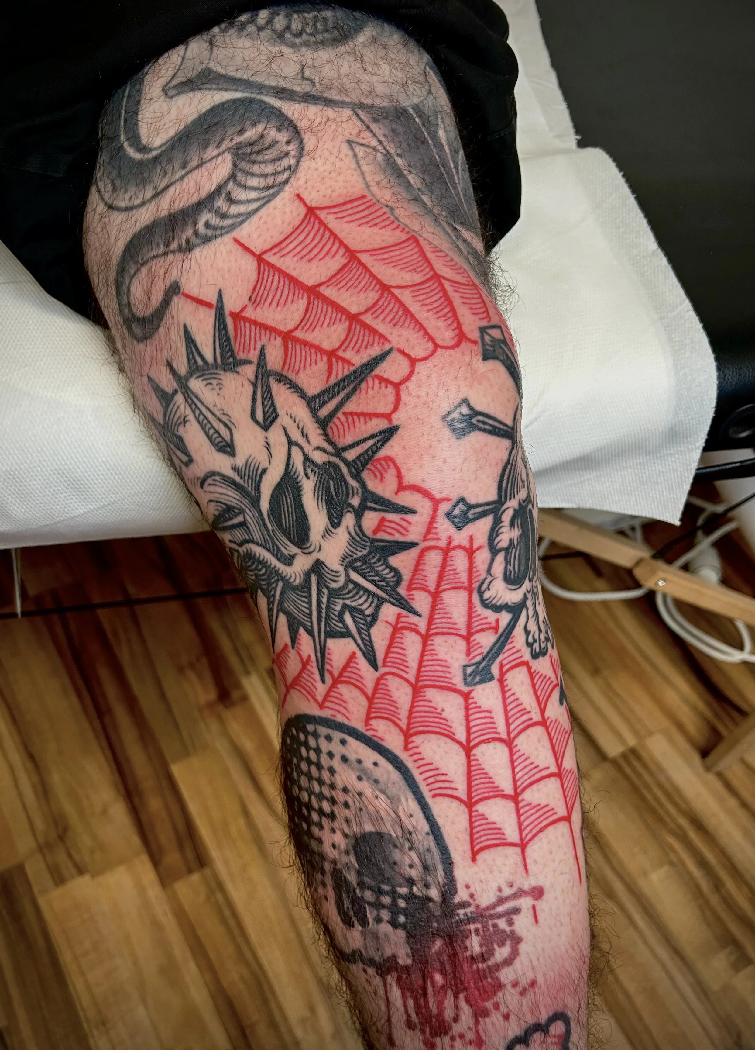 Tatouage d'une araignée rouge sur une jambe, entouré d'autres tatouages en noir, dont un crâne et des éléments décoratifs.