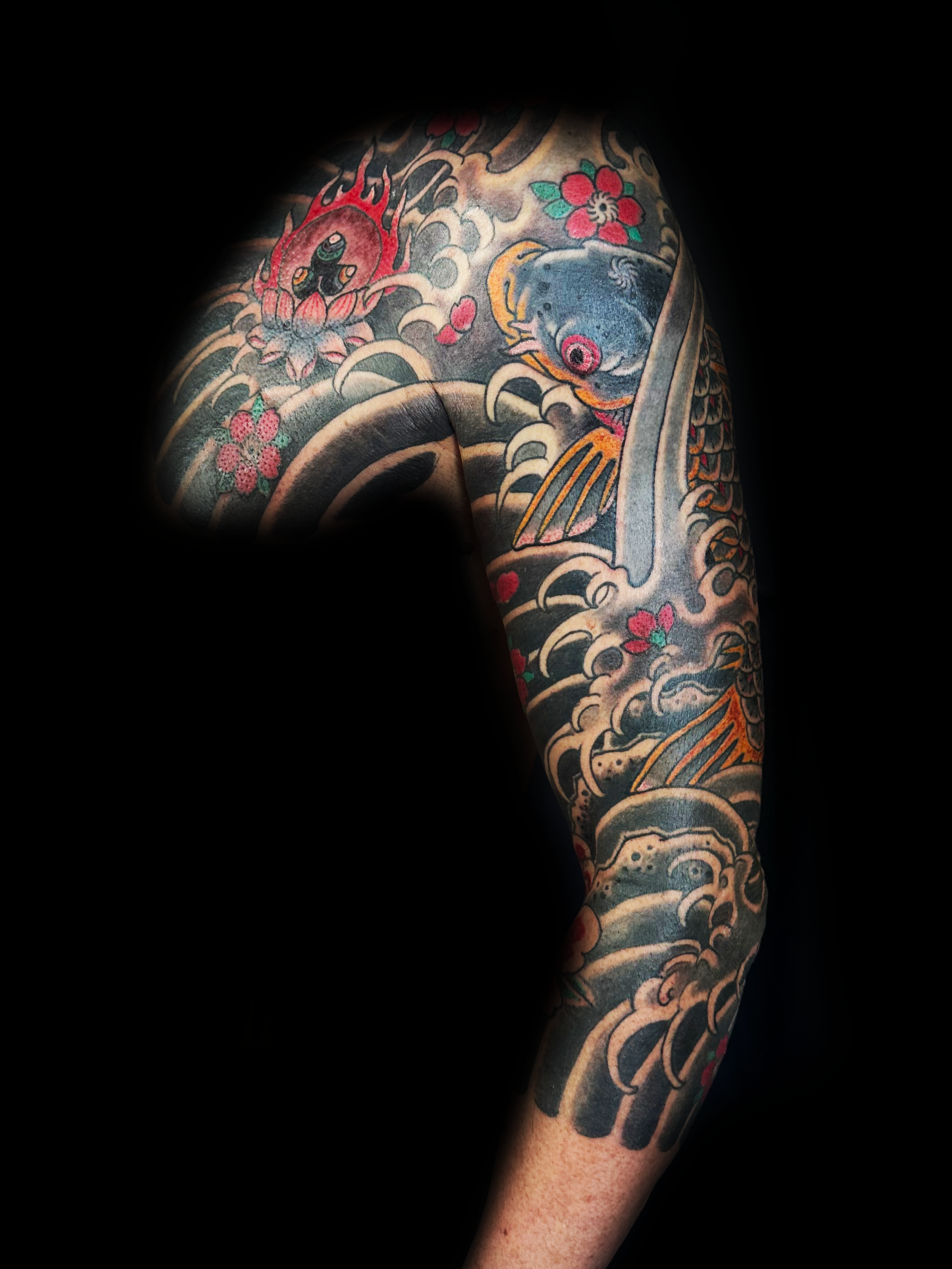 Tatouages de style traditionnel japonais sur le bras, représentant un poisson, des fleurs et d'autres motifs, avec un fond noir.