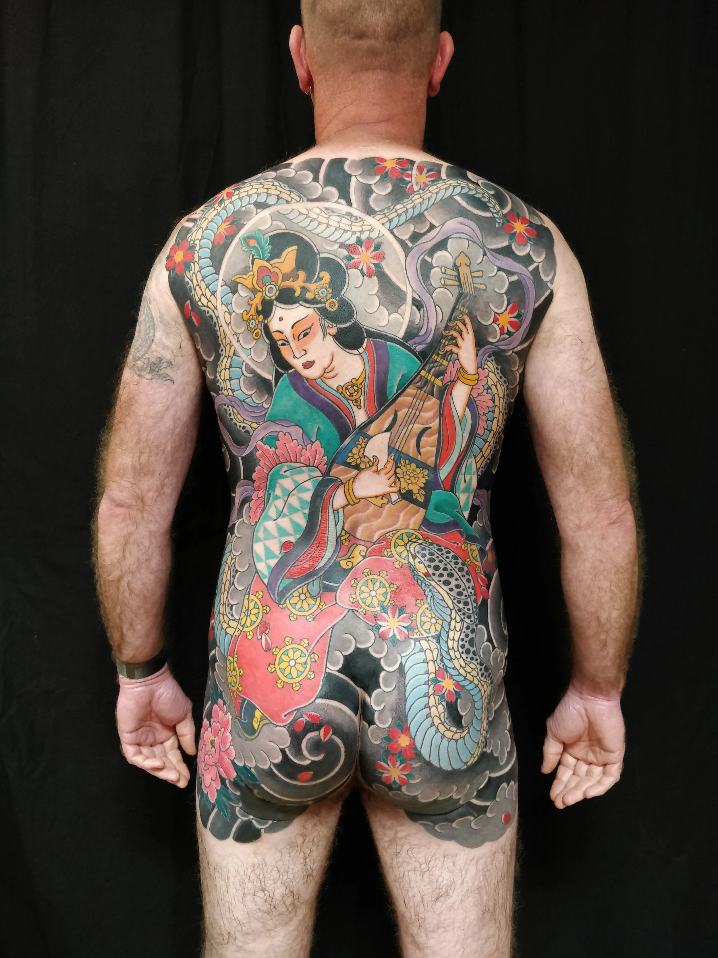 Tatouage coloré d'une femme jouant de la guitare, entourée de fleurs et de nuages, couvrant la majorité du dos et des jambes d'un homme.