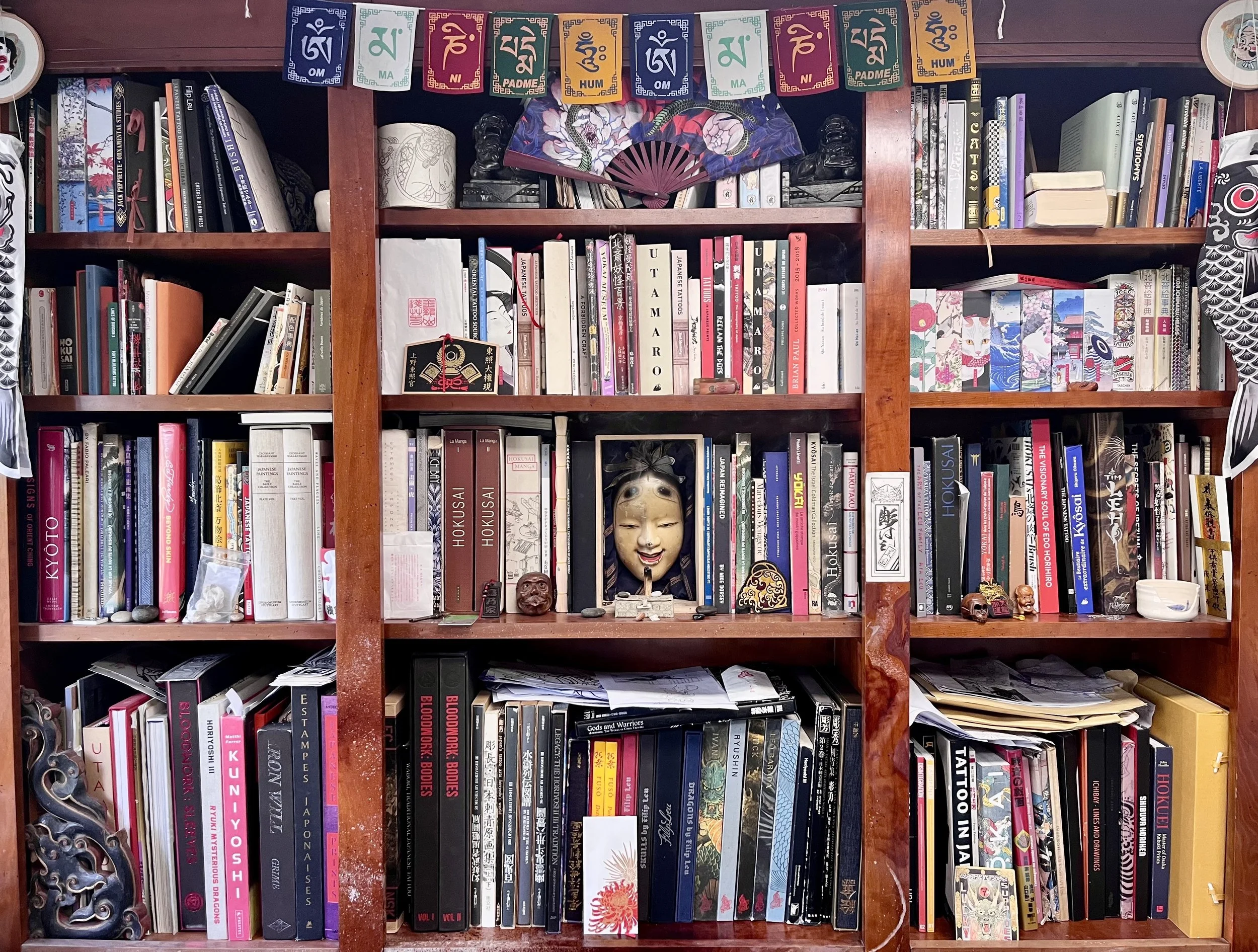 Une étagère en bois remplie de livres de différentes couleurs et tailles, avec des objets décoratifs comme des masques et une fanfare pliée, et des guirlandes de drapeaux colorés en haut de l'étagère.