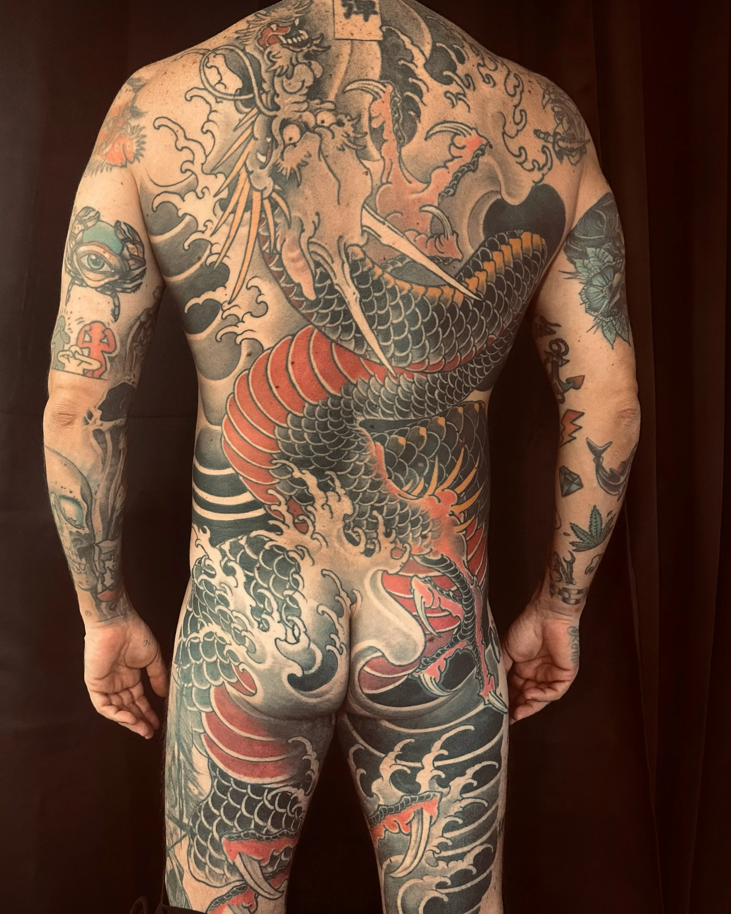 Tatouage coloré de dragon japonais couvrant tout le dos et les bras d'un homme. Le dragon a des détails en orange, noir et blanc, avec des vagues et des nuages en arrière-plan.