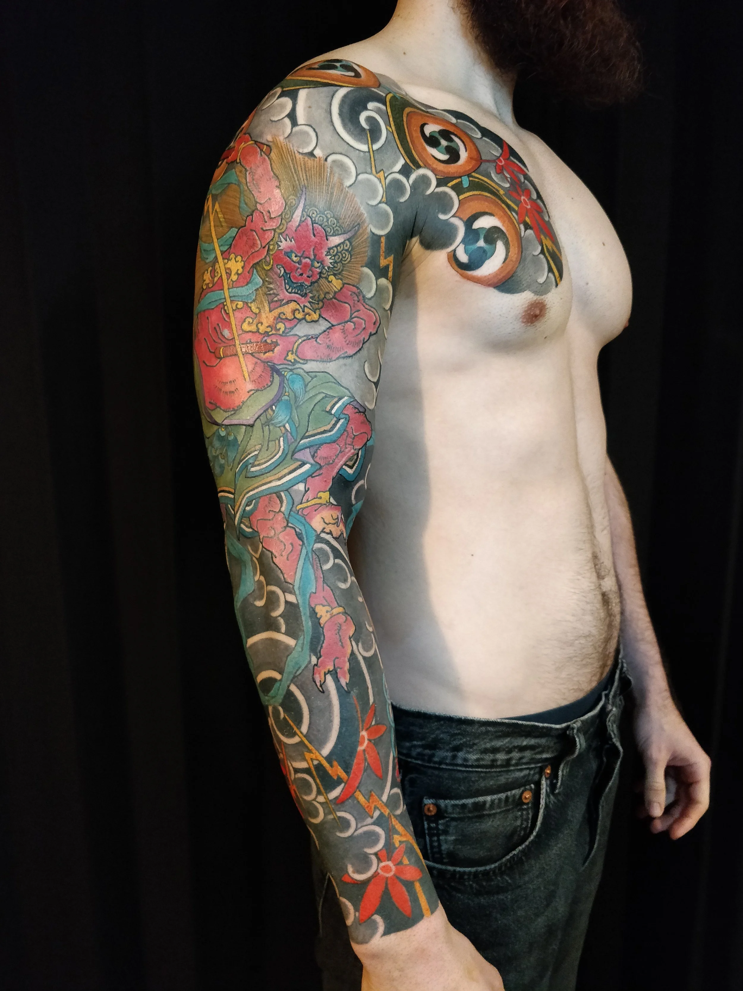 Un homme avec un tatouage coloré couvrant son épaule et son bras, représentant un dragon et d'autres motifs traditionnels, vêtu d'un t-shirt noir et de jeans, contre un fond noir.