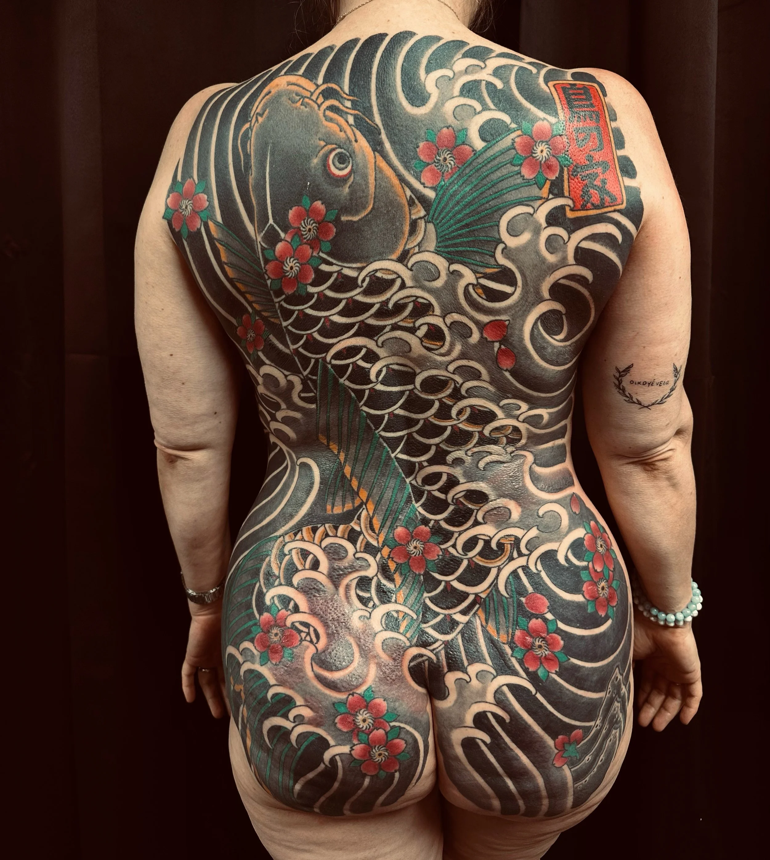 Tatouage japonais traditionnel sur le dos d'une personne, représentant une vague avec un poisson, des fleurs rouges, un éventail et un tibis.