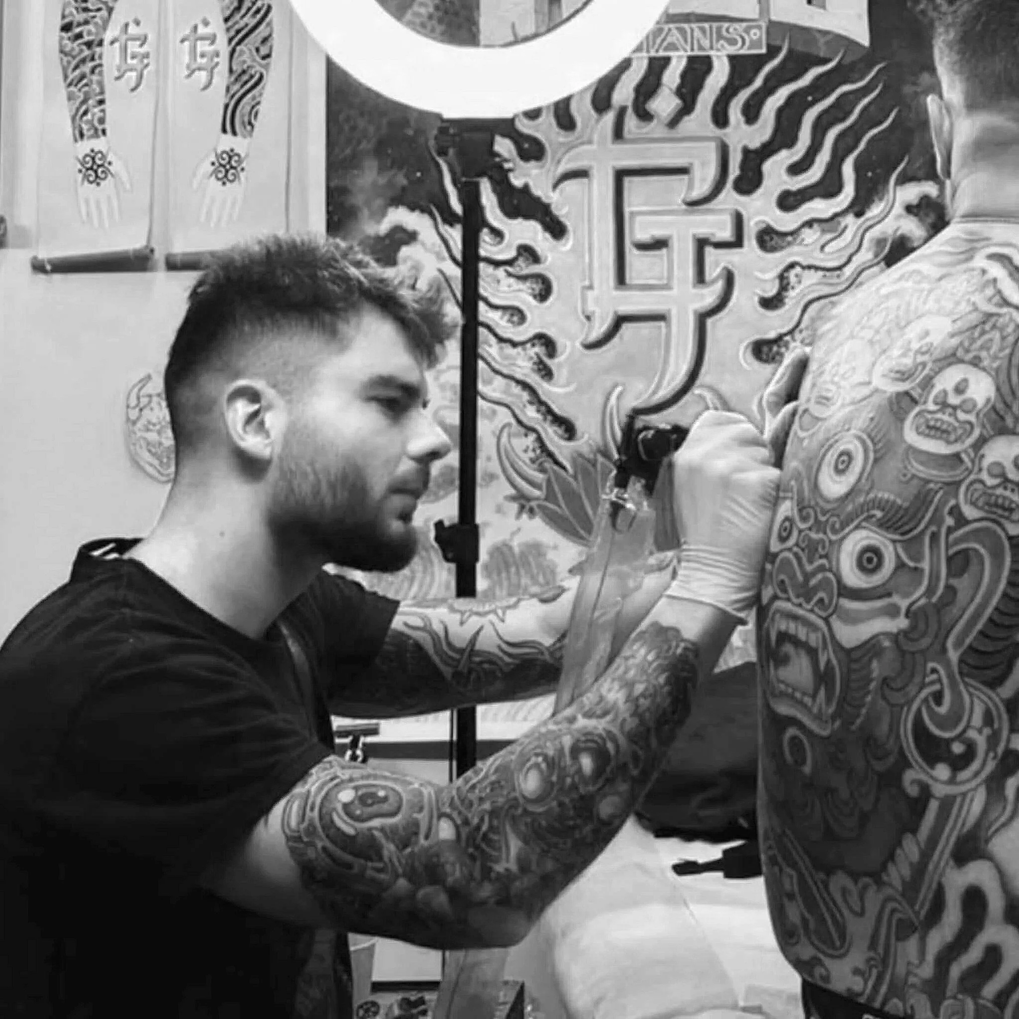 Photographie en noir et blanc montrant une personne avec une coiffure courte et une barbe, assise devant un fond décoratif avec des motifs et des drapeaux.